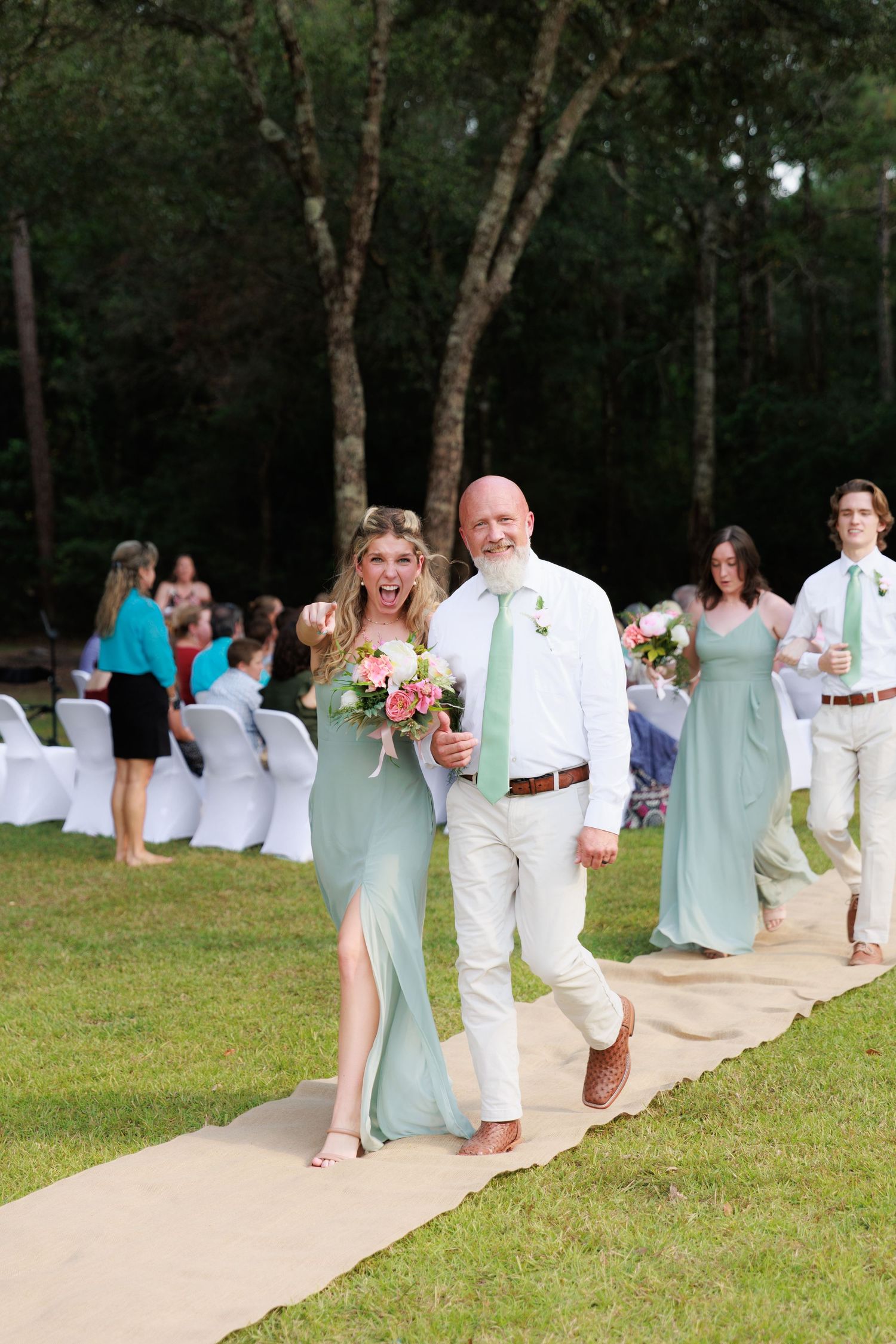 Wedding party members in mint green and white attire walking down an outdoor ceremony aisle.