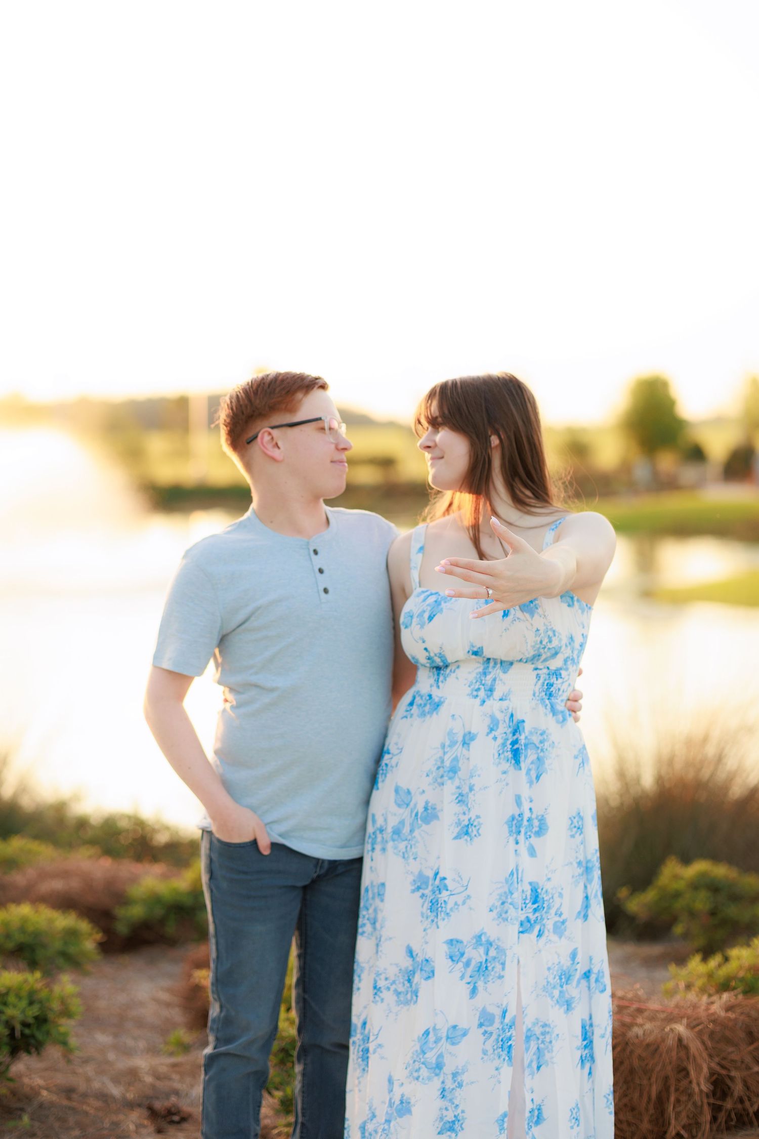 Couple sharing a tender moment outdoors near a lake at golden hour, woman in floral dress, man in gray shirt.