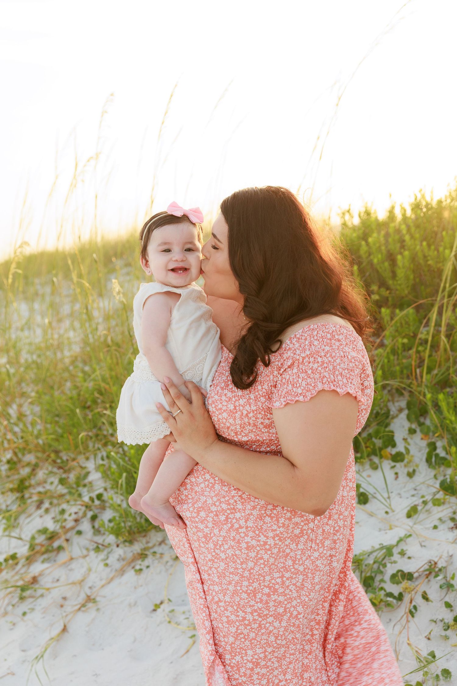 A mother in a floral dress kisses her baby girl on a sandy beach with sea oats in the background.