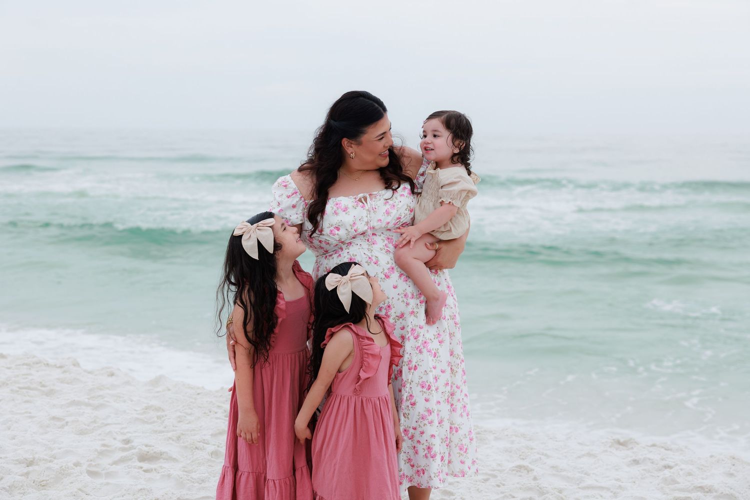 A mother in a floral dress holds a toddler while two girls in pink dresses stand beside her on a beach.