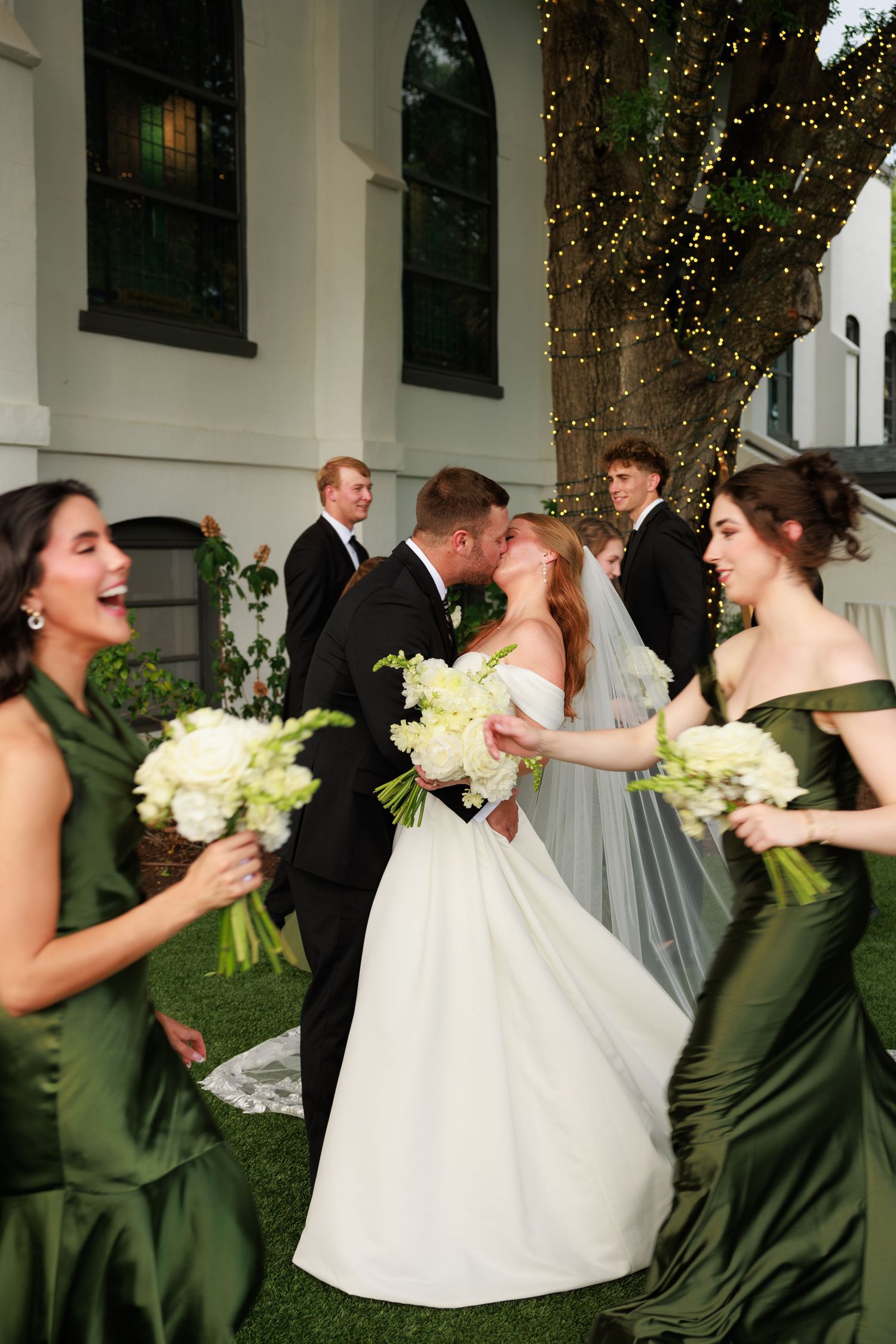 Bride and groom share a kiss surrounded by wedding party in green gowns and black tuxedos outdoors near a white building.