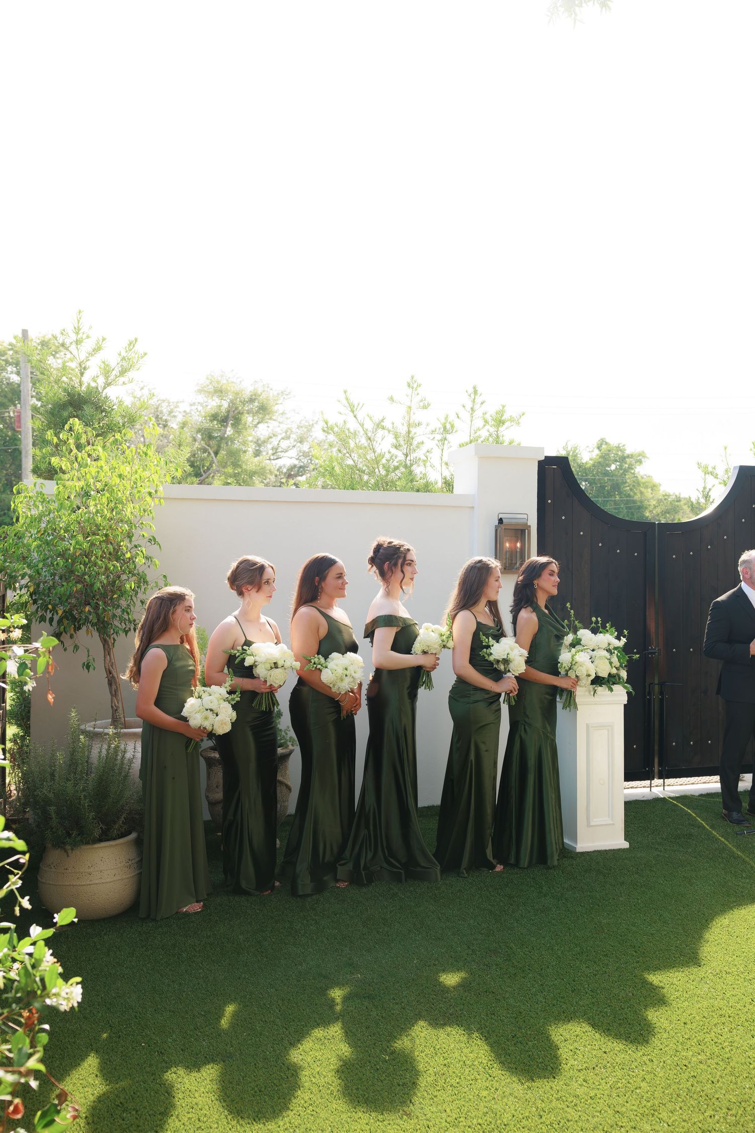 Bridesmaids in elegant green gowns holding white floral bouquets stand in a row at an outdoor wedding ceremony.
