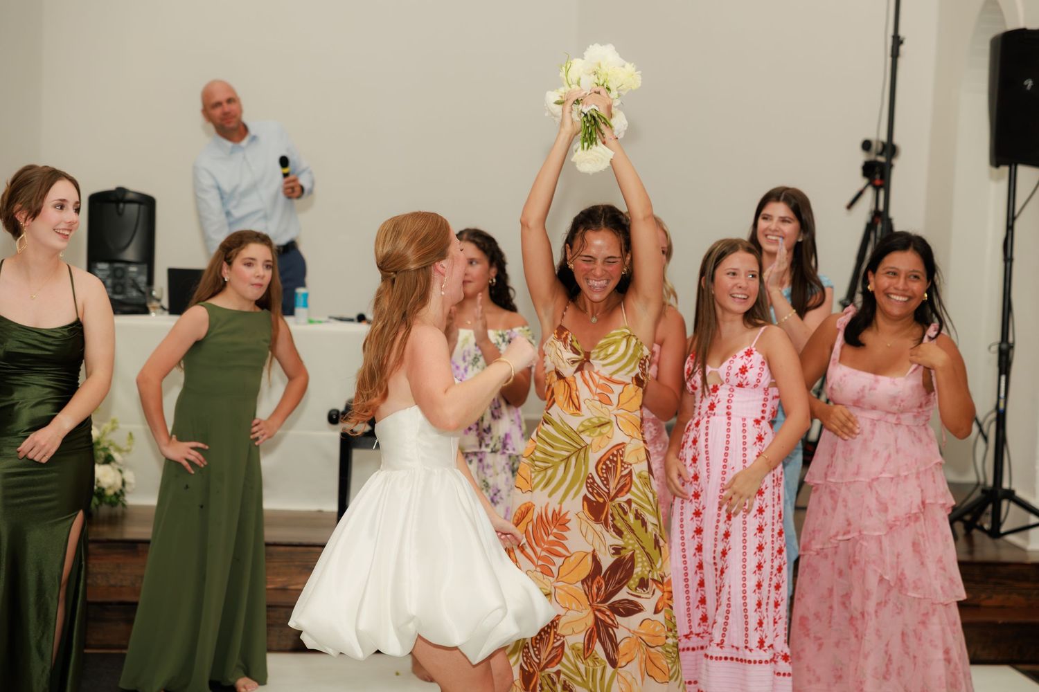 Bride watches as woman in floral dress catches wedding bouquet toss surrounded by smiling bridesmaids.