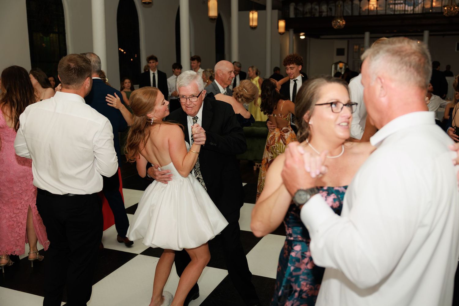 Guests dancing joyfully at an elegant wedding reception on a black and white checkered dance floor.