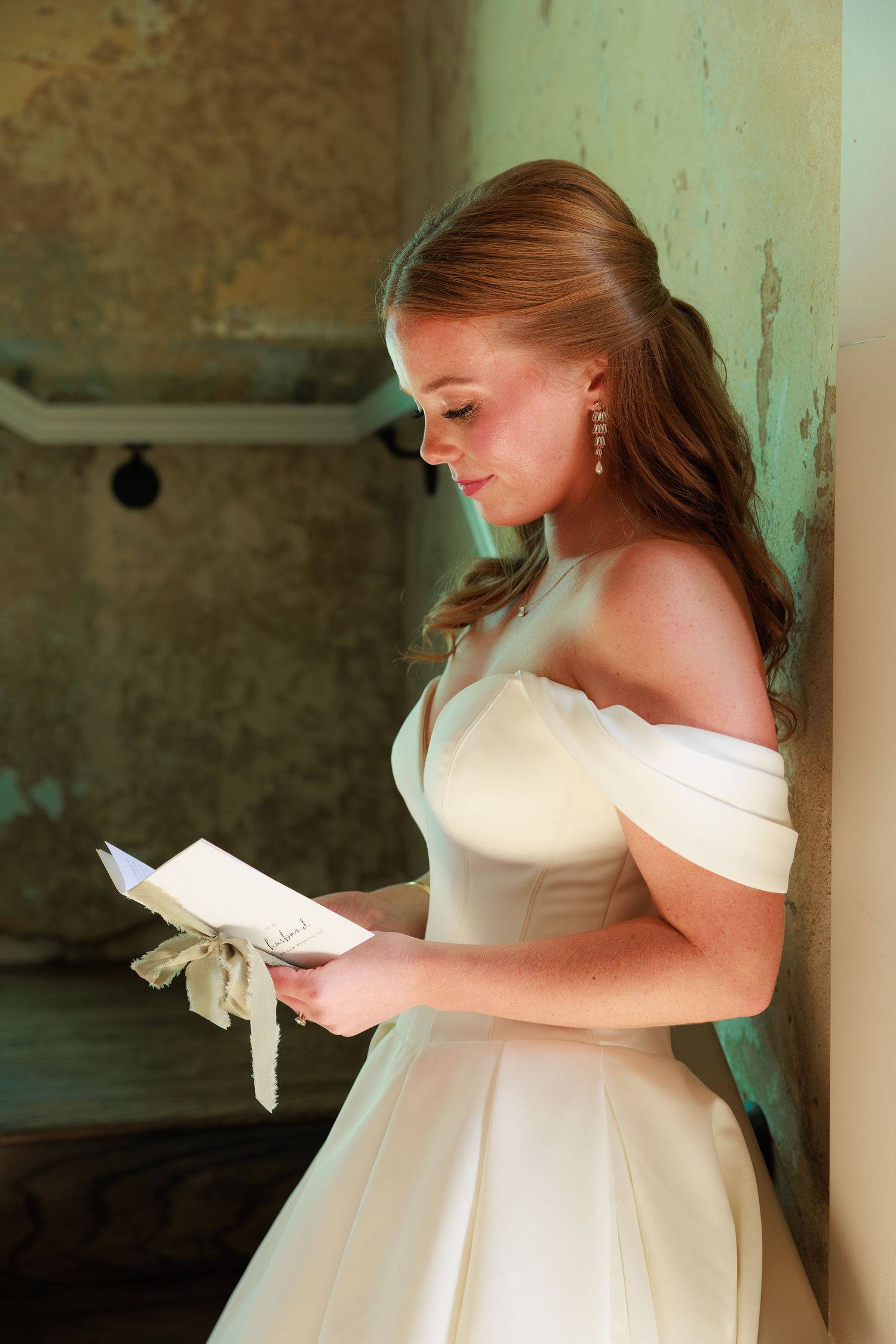 Bride in off-shoulder white gown reads a heartfelt letter, leaning against a vintage wall.