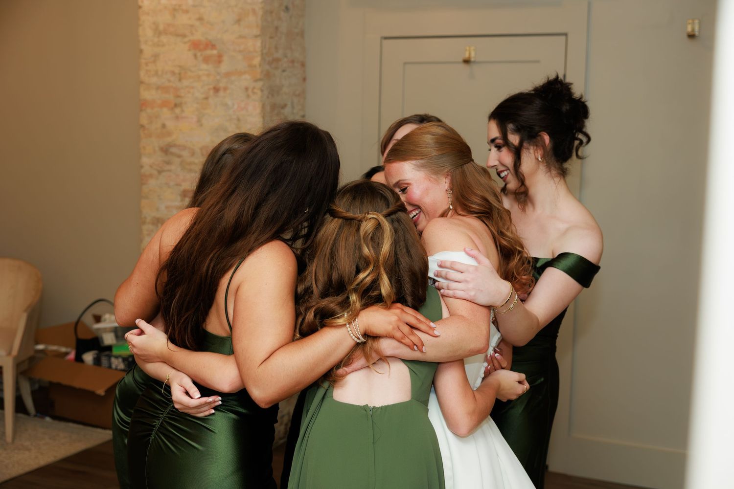 Bride in white gown shares emotional group hug with bridesmaids in green satin dresses before wedding ceremony.