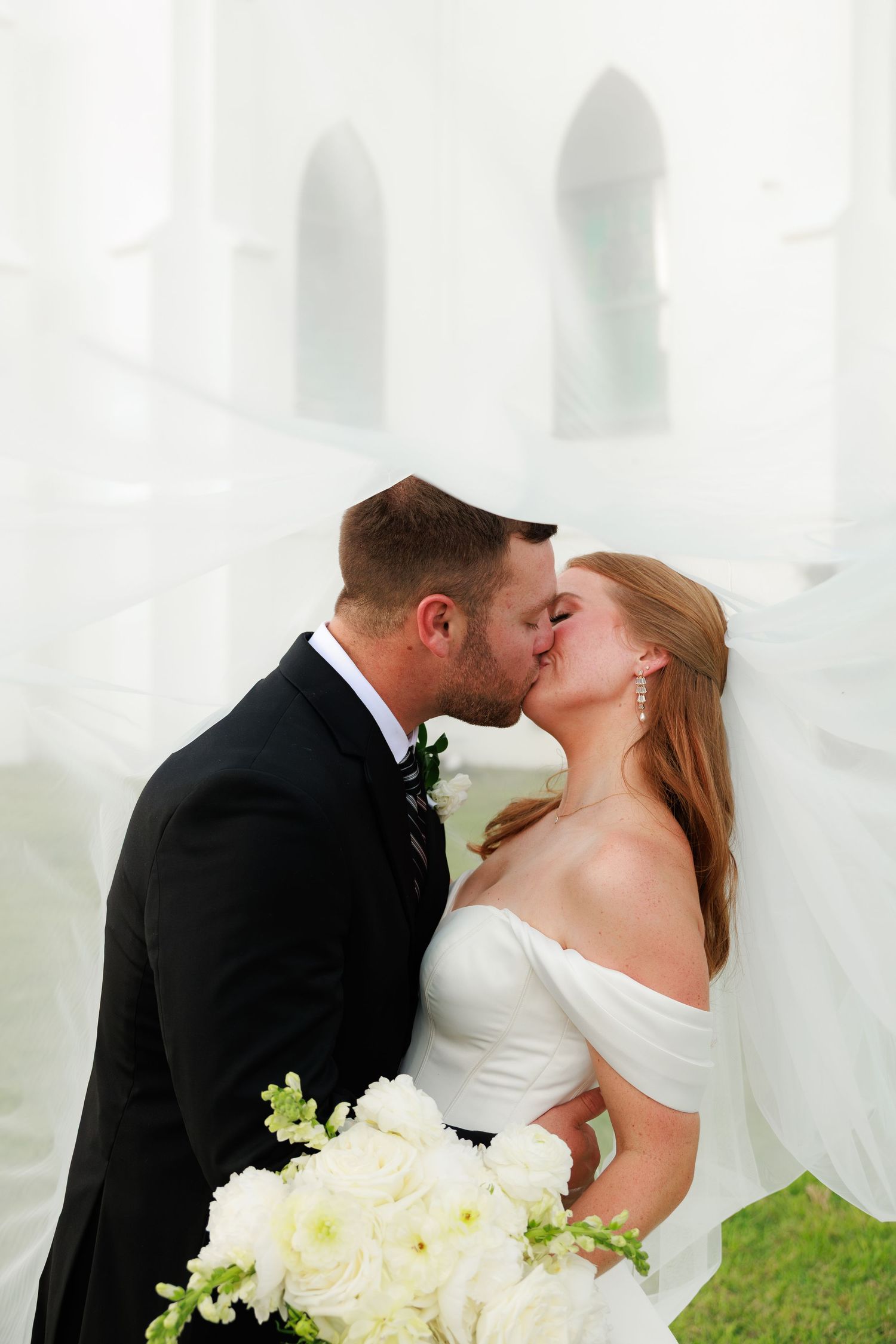 Bride and groom share a kiss framed by flowing white veil with white floral bouquet outdoors.
