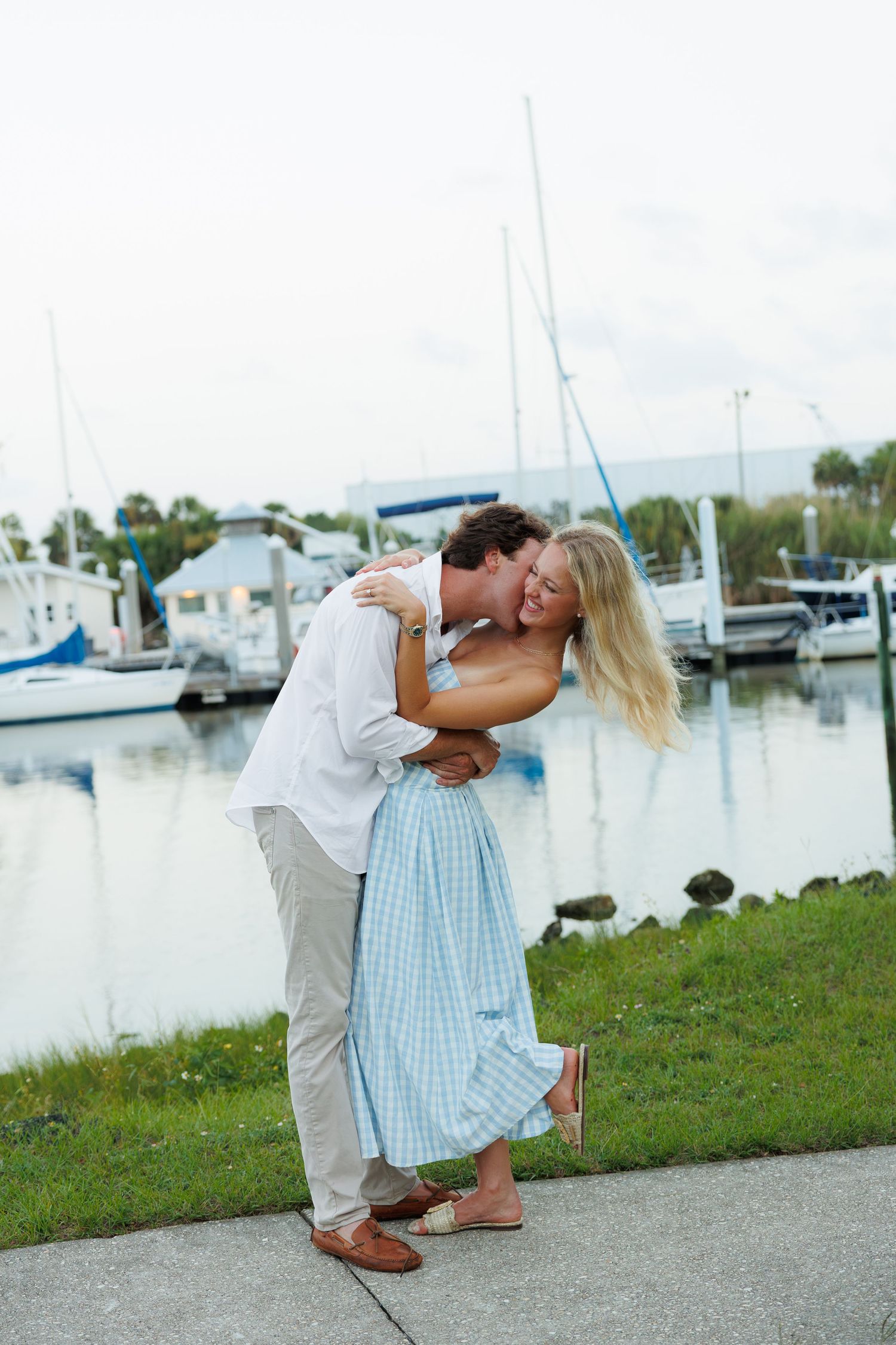 Couple embracing and kissing near a marina with sailboats, woman in blue dress, man in white shirt.