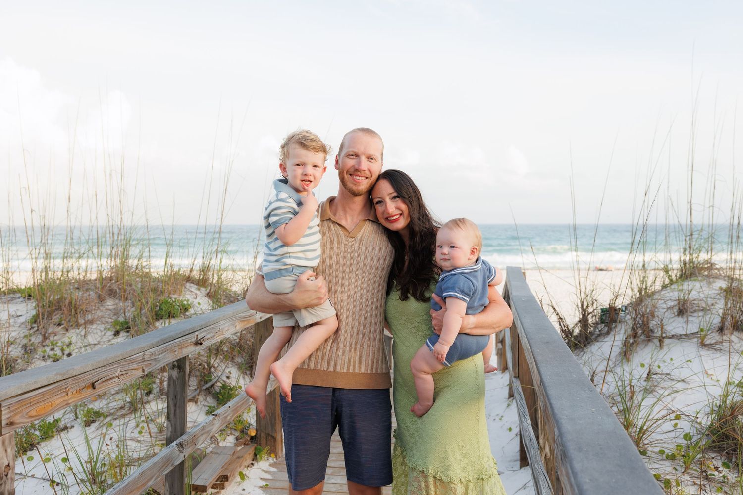 Family of four smiling on a beach boardwalk, parents holding two young children with ocean and sea oats in background.