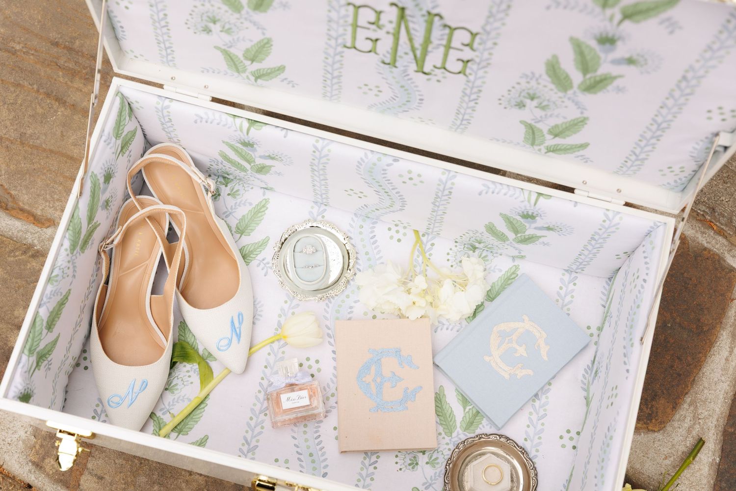 An open decorative box with monogram lining displaying nude ballet flats, perfume, blue stationery cards, and floral accents.