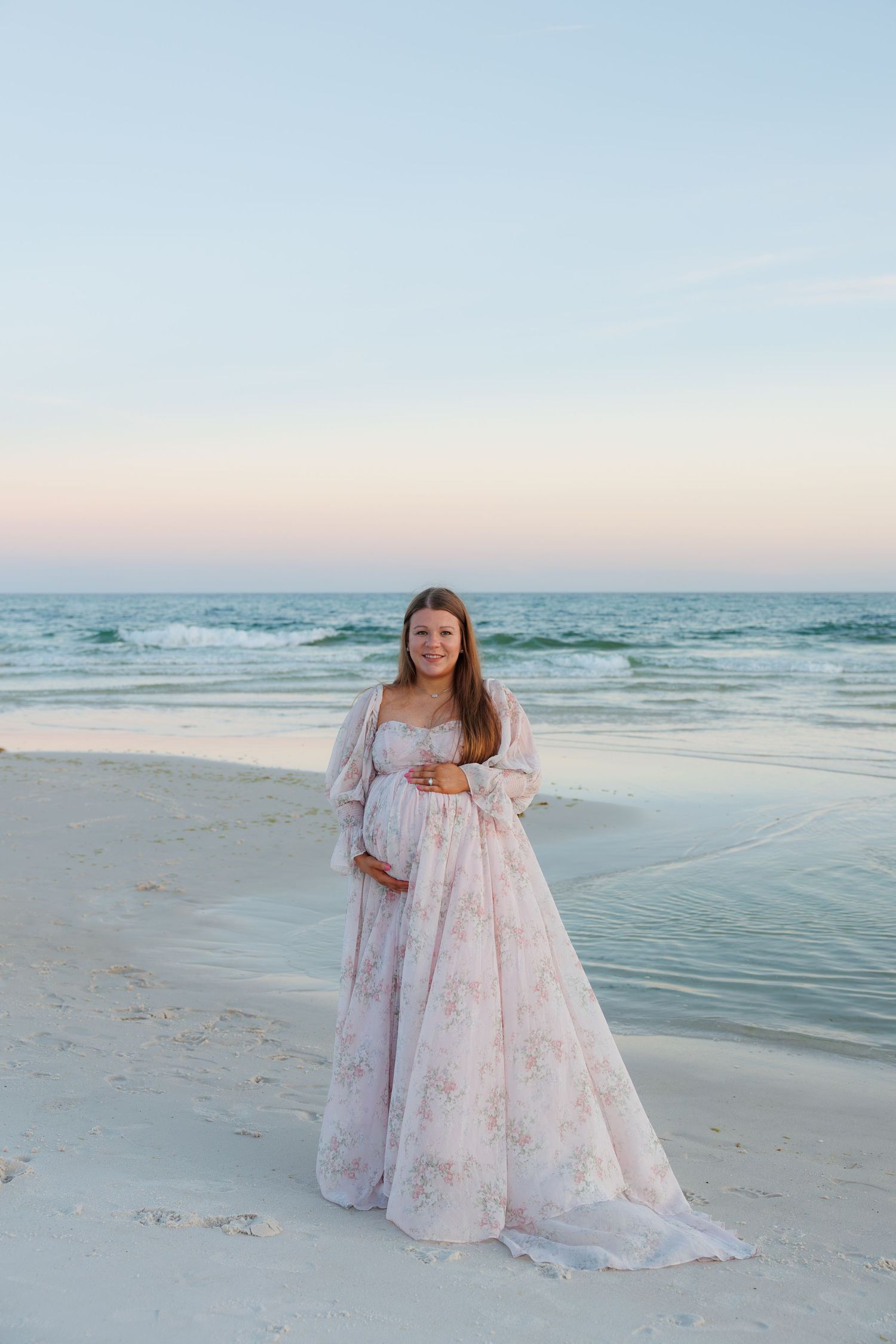 Pregnant woman in flowing floral dress standing on white sand beach at sunset by ocean.