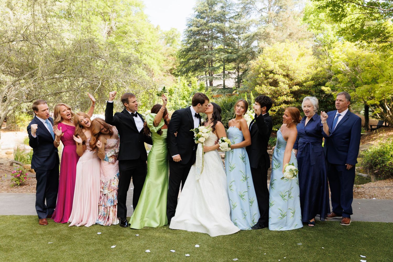 Wedding party poses outdoors as bride and groom kiss, surrounded by guests in colorful gowns and tuxedos in a lush garden.