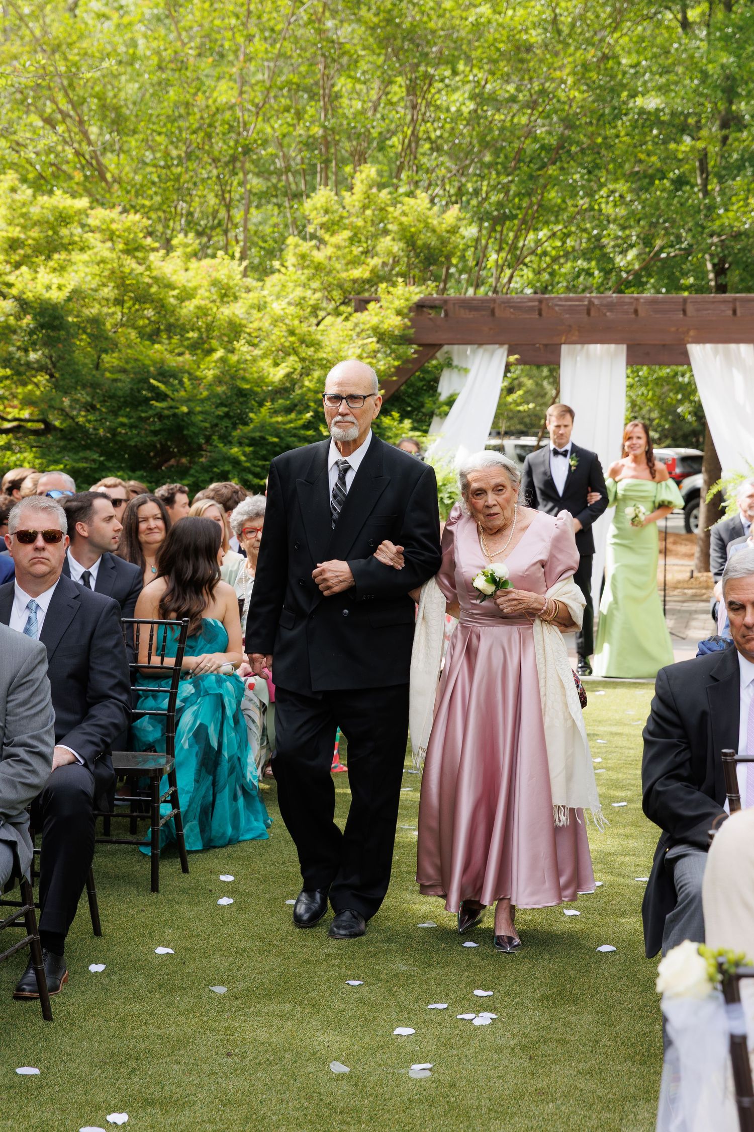 Elderly couple walks down outdoor wedding aisle, woman in pink dress holding flowers, surrounded by seated guests on green lawn.