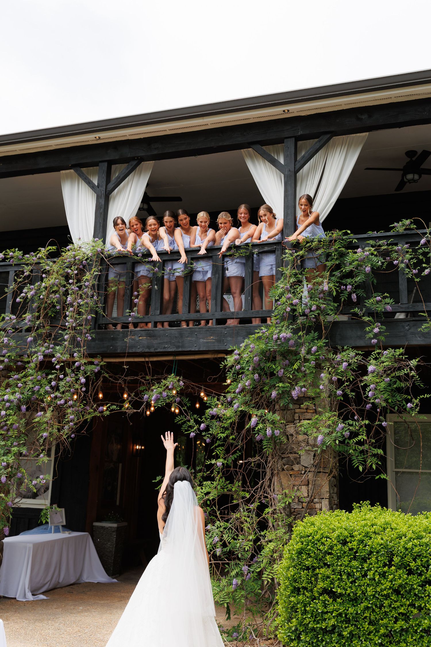 Bride in white gown waves up to bridesmaids in grey dresses on a vine-covered balcony at a wedding venue.