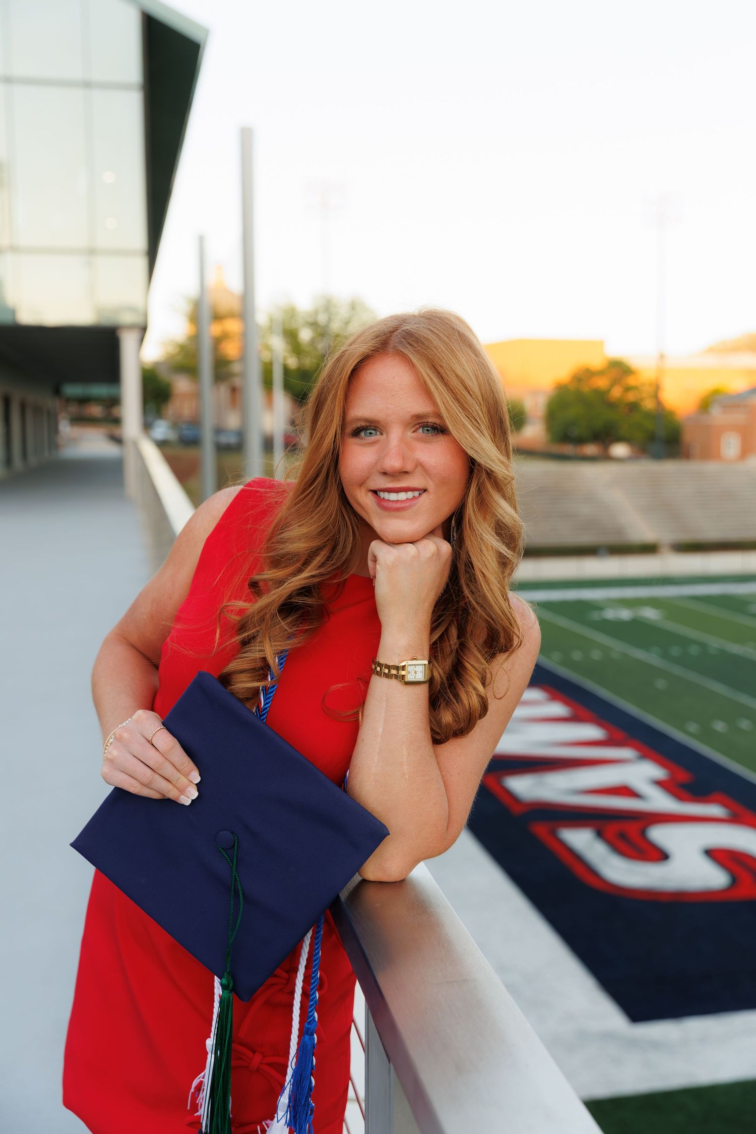 Smiling young woman in red dress holding navy graduation cap on stadium balcony at golden hour.