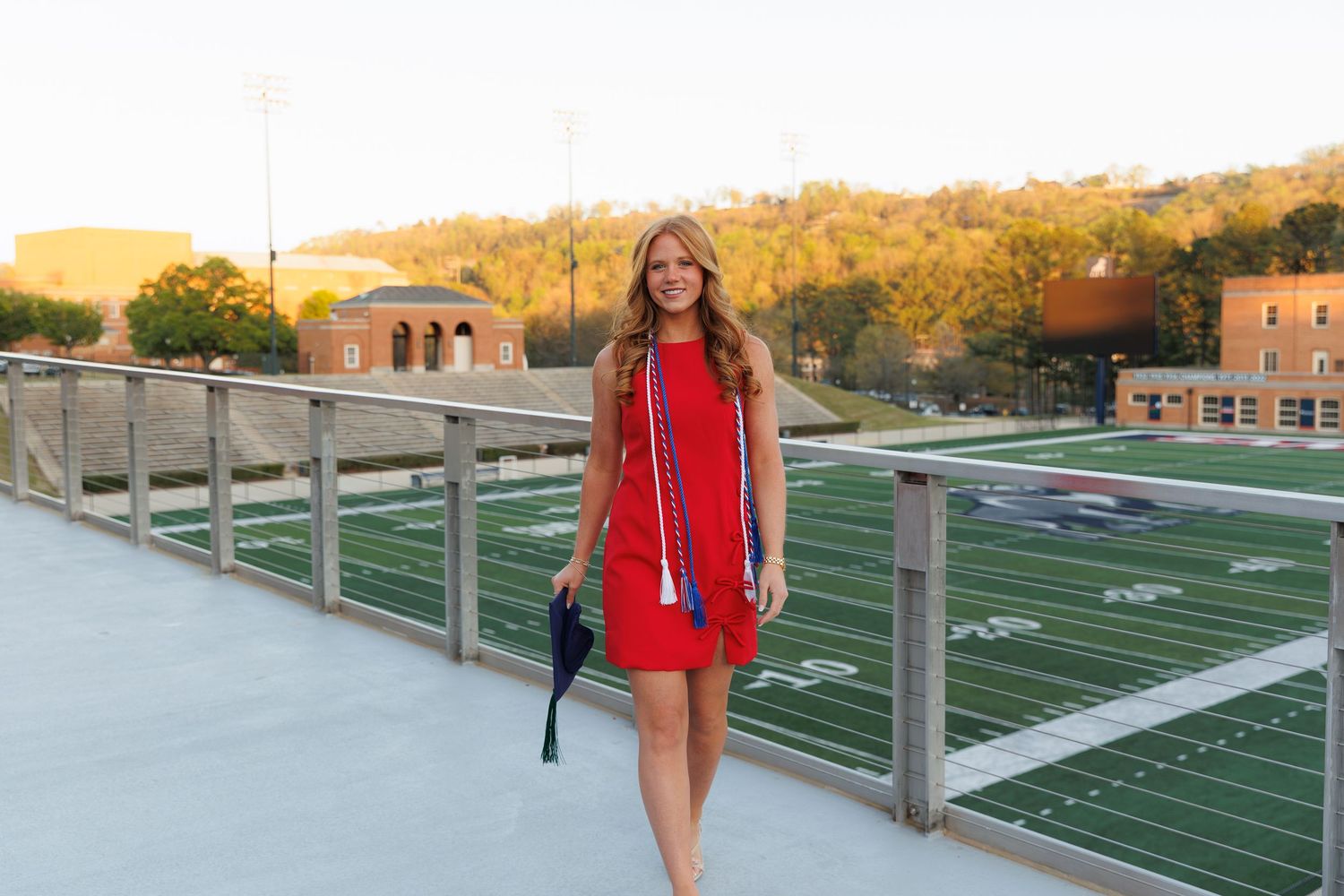 Young woman in red dress holding graduation cap on stadium bridge, celebrating college graduation outdoors.