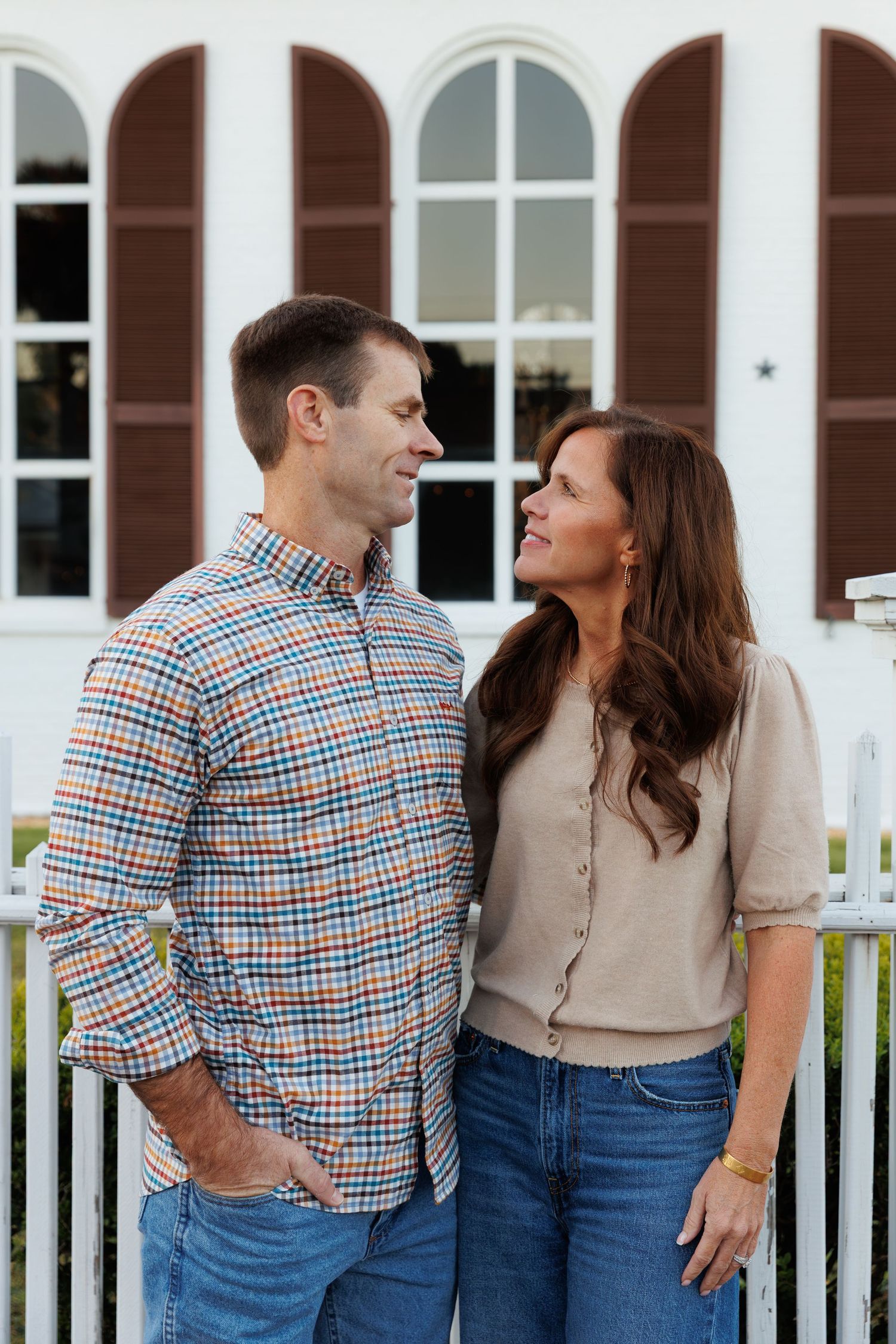Smiling couple standing together outdoors by a white fence, man in plaid shirt and woman in beige cardigan.
