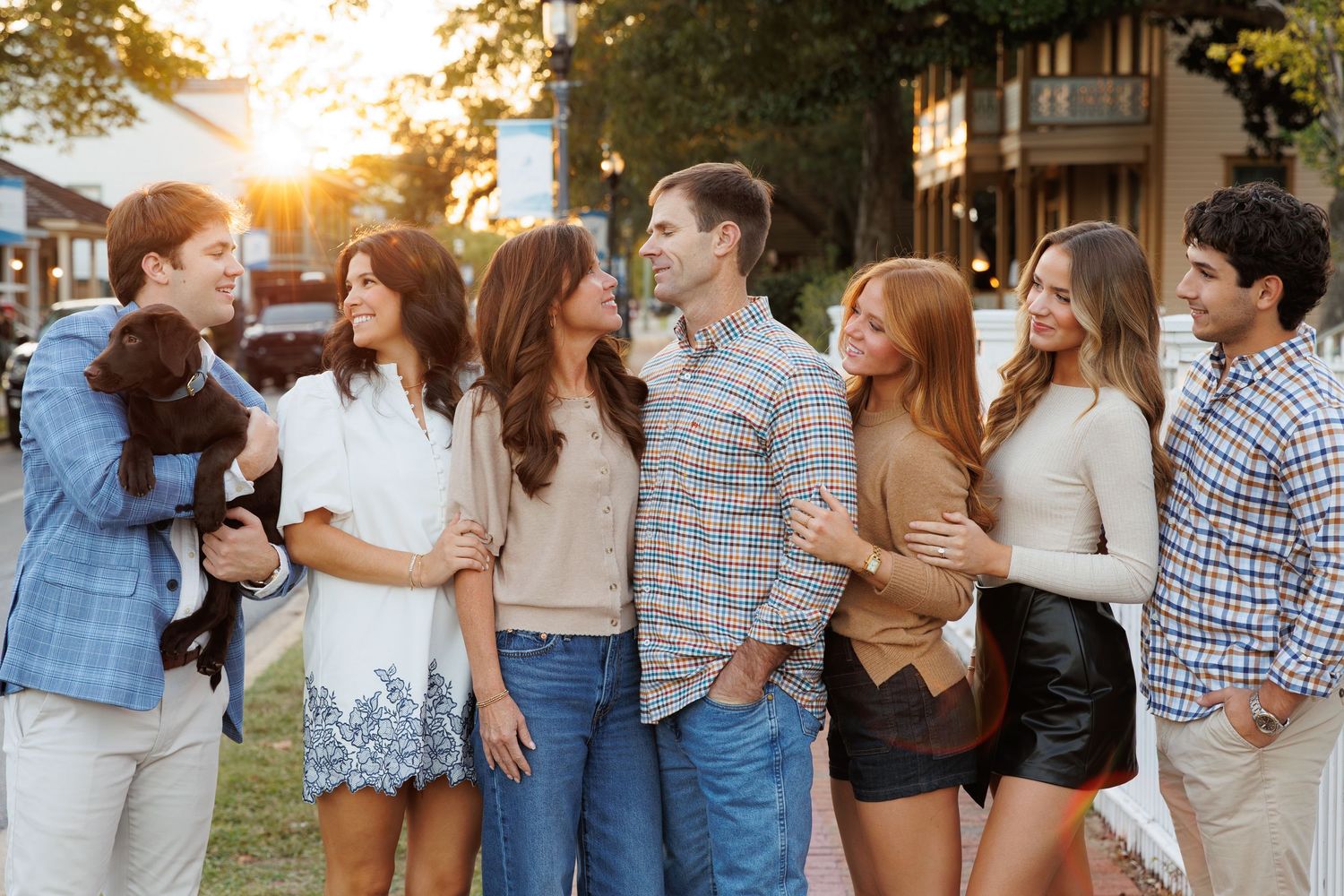 Group of seven young adults socializing outdoors at sunset, casually dressed, one holding a small dog.