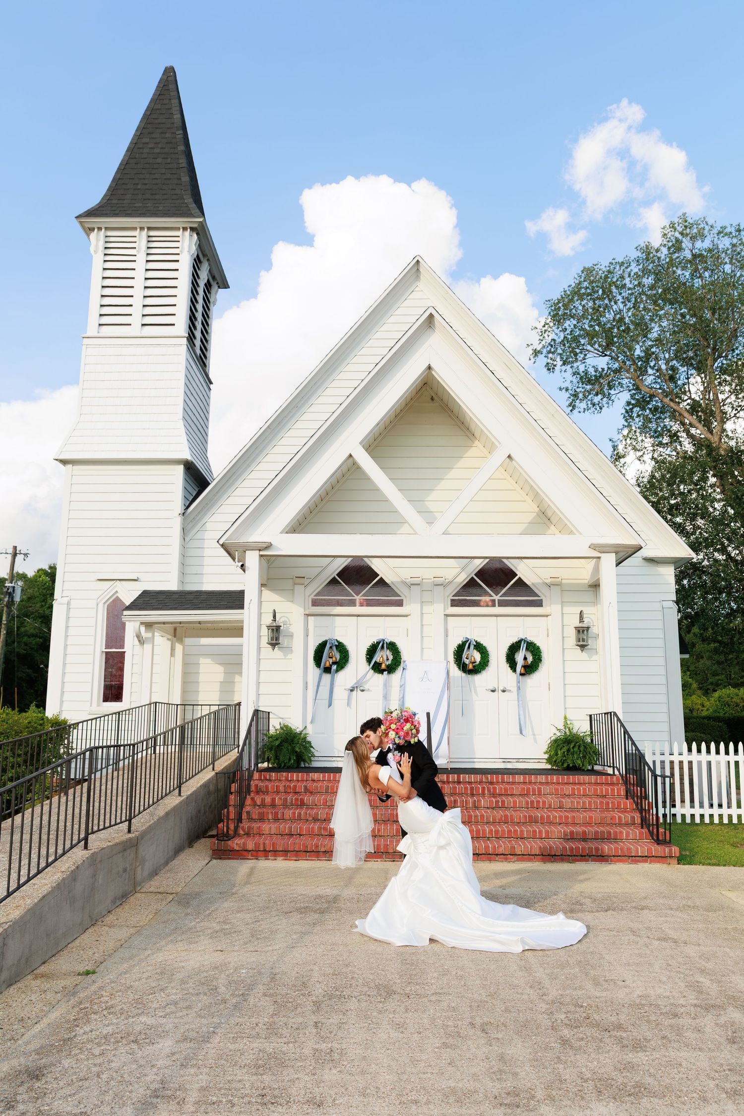 Wedding couple poses in front of a charming white chapel with Christmas wreaths on a sunny day.
