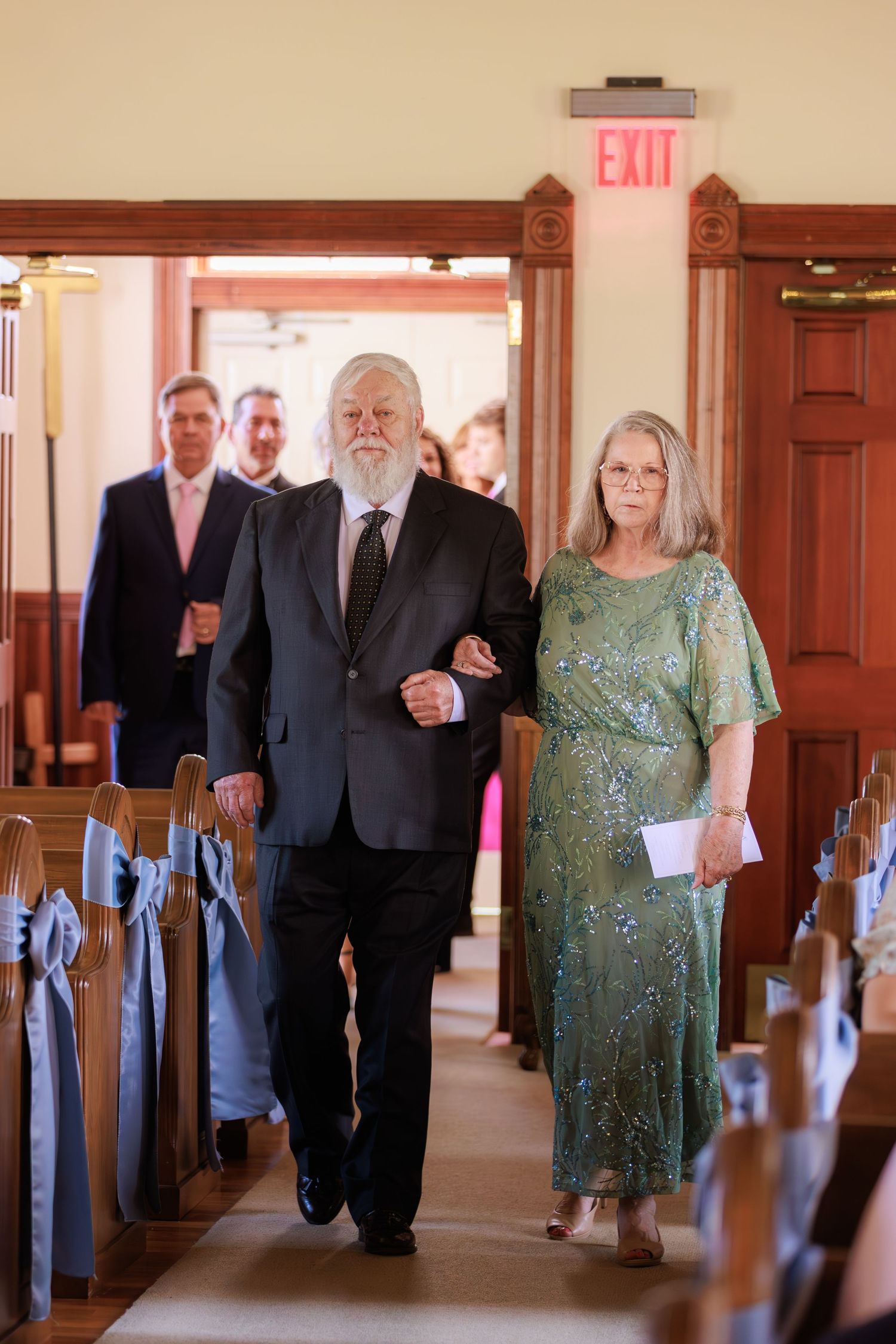 Wedding guests walk down the aisle of a traditional church with wooden pews and an illuminated exit sign.