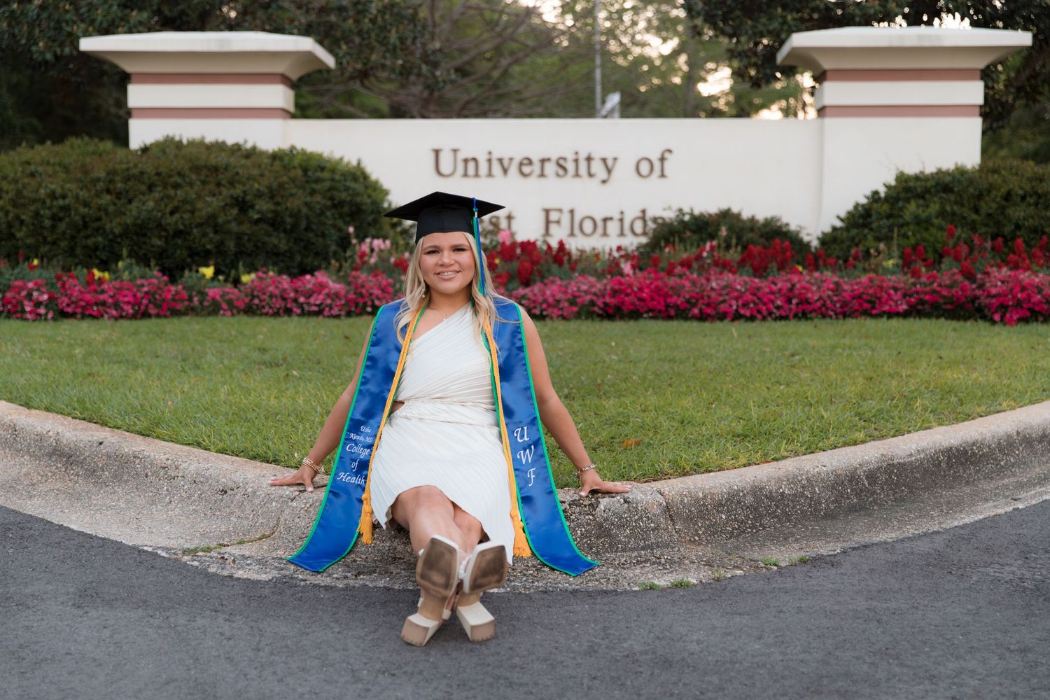 Graduate in academic regalia sits in front of University of Florida entrance sign with red flowers.