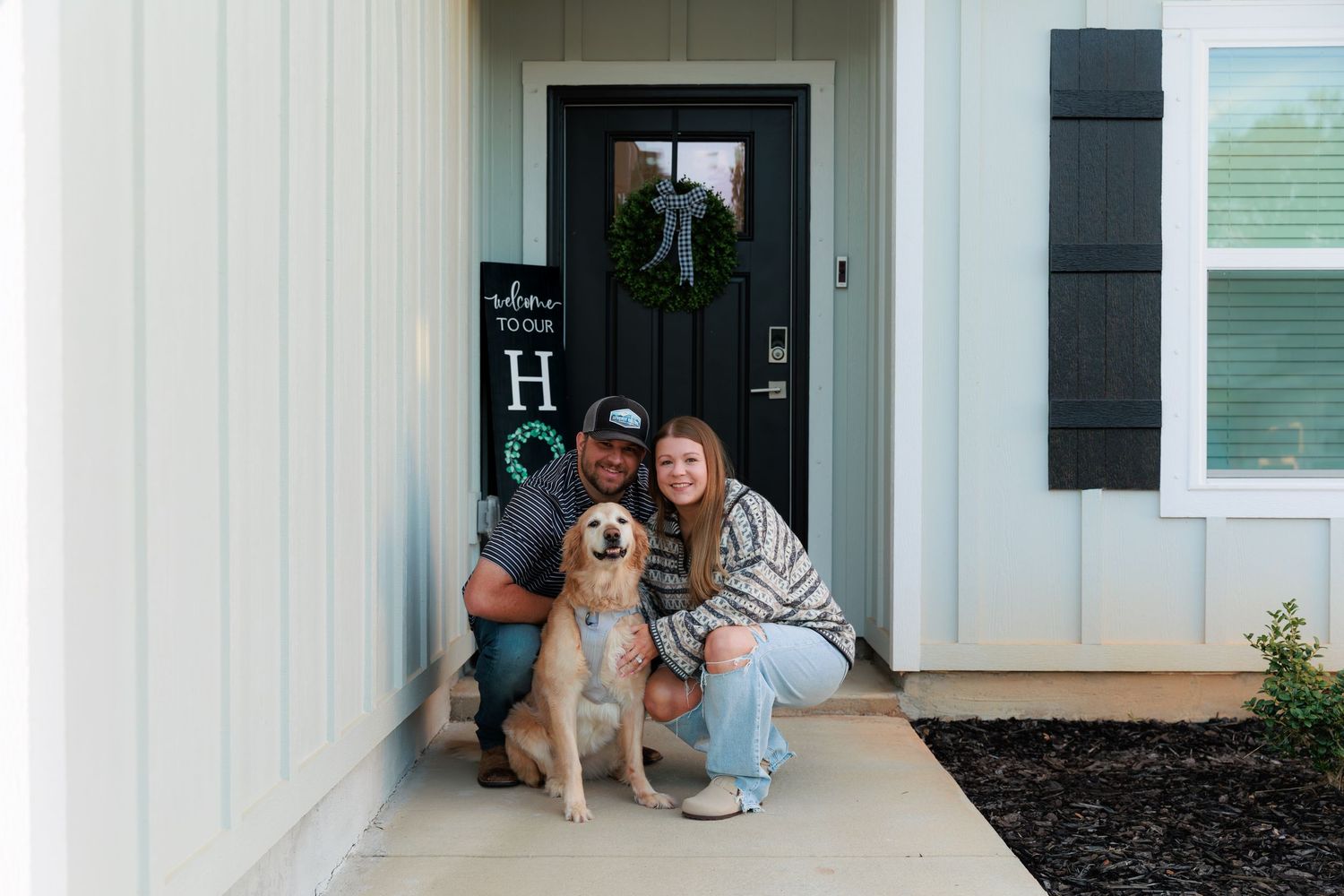 A couple sits with their golden retriever on their front porch by a white farmhouse with black shutters and a wreath on the door.
