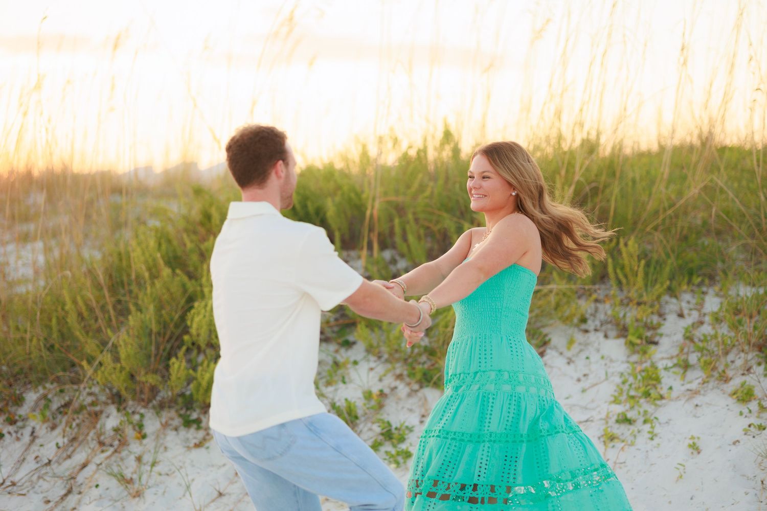 A couple dances playfully together on a sandy beach at sunset with ocean waves in the background.
