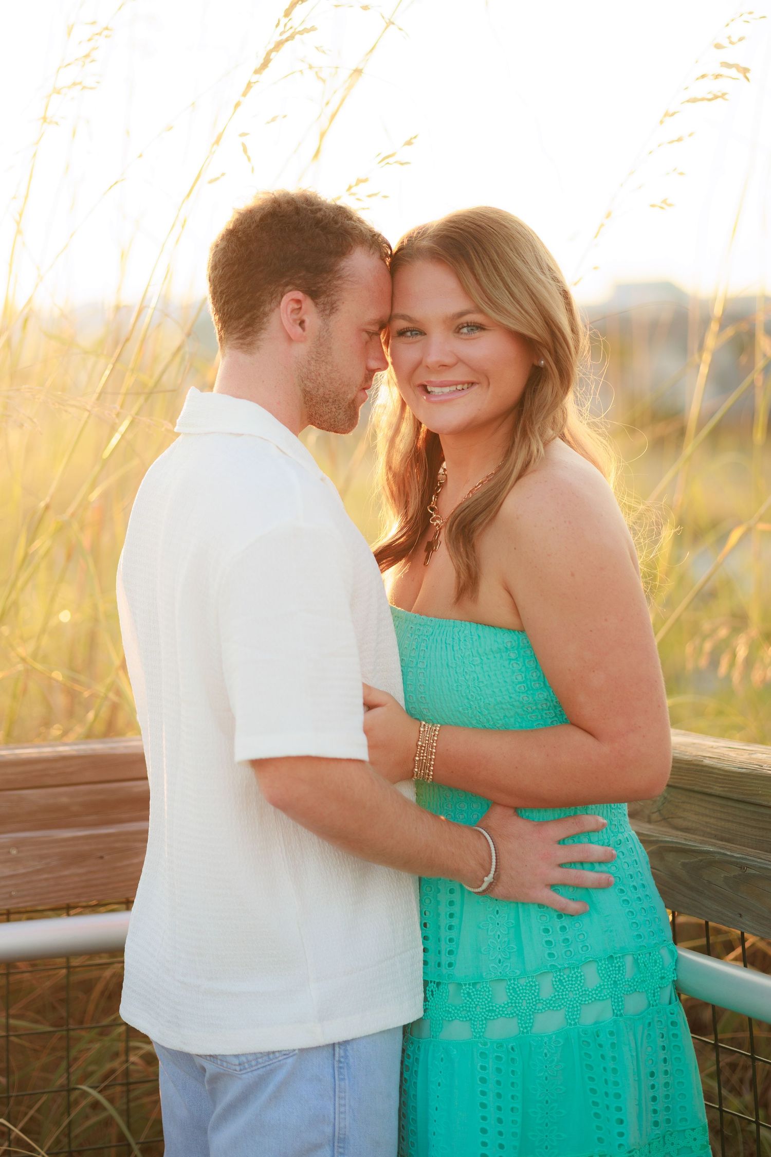 Romantic couple embraces on beach boardwalk at sunset, woman in turquoise dress and man in white shirt.