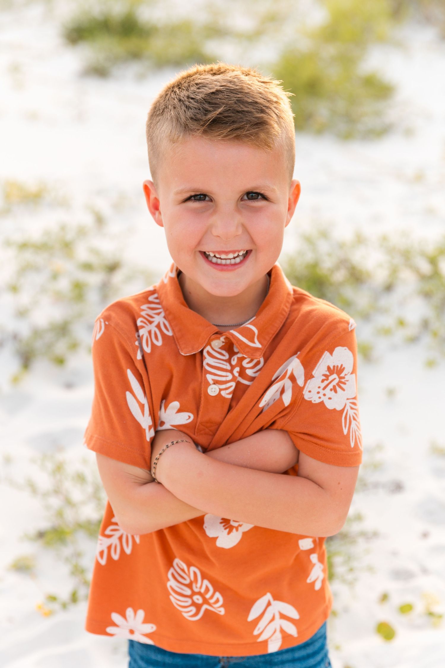 Young boy in orange floral shirt smiles confidently with arms crossed.