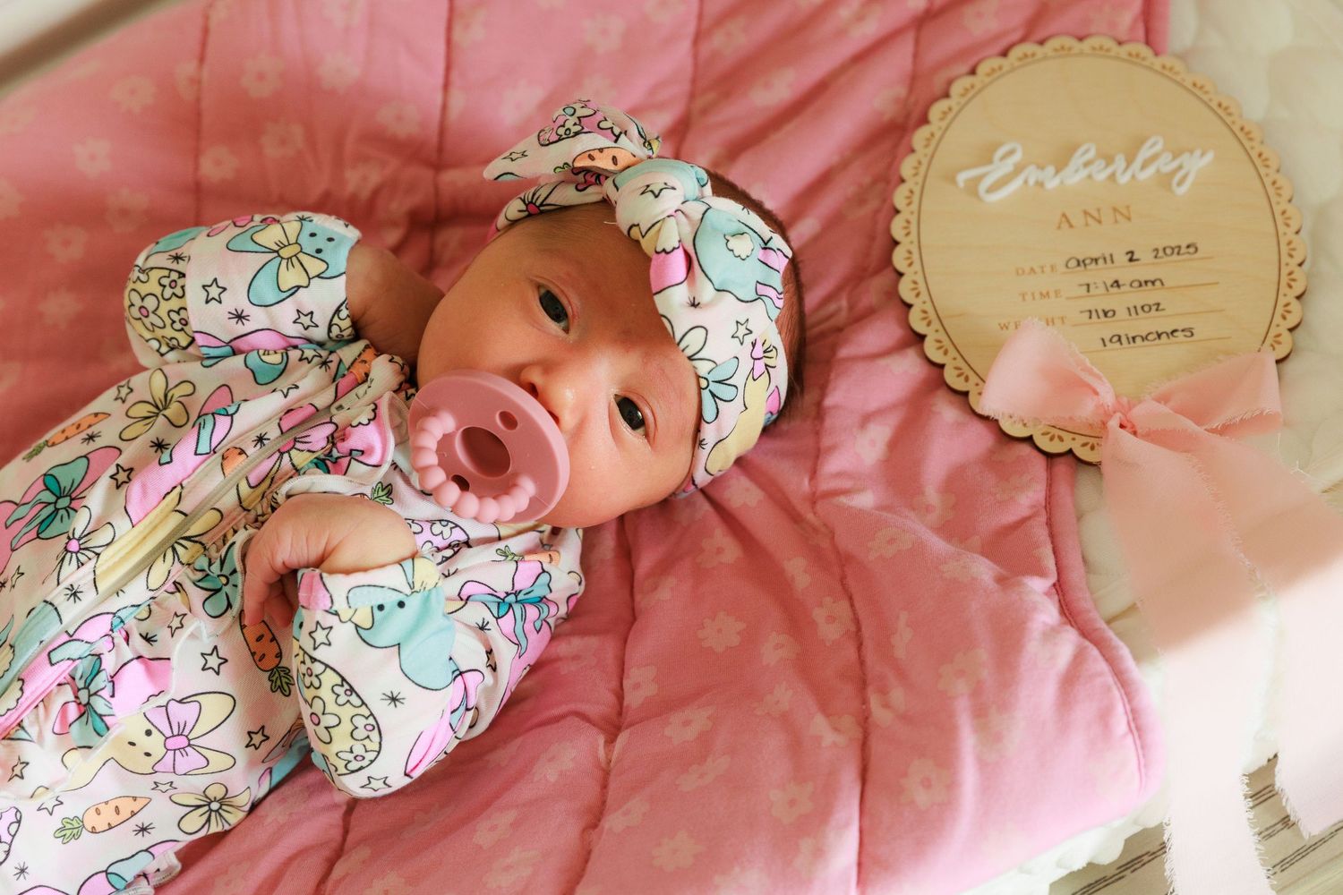 A newborn baby lying on pink bedding with floral accessories and decorative elements in a lifestyle photography setting.