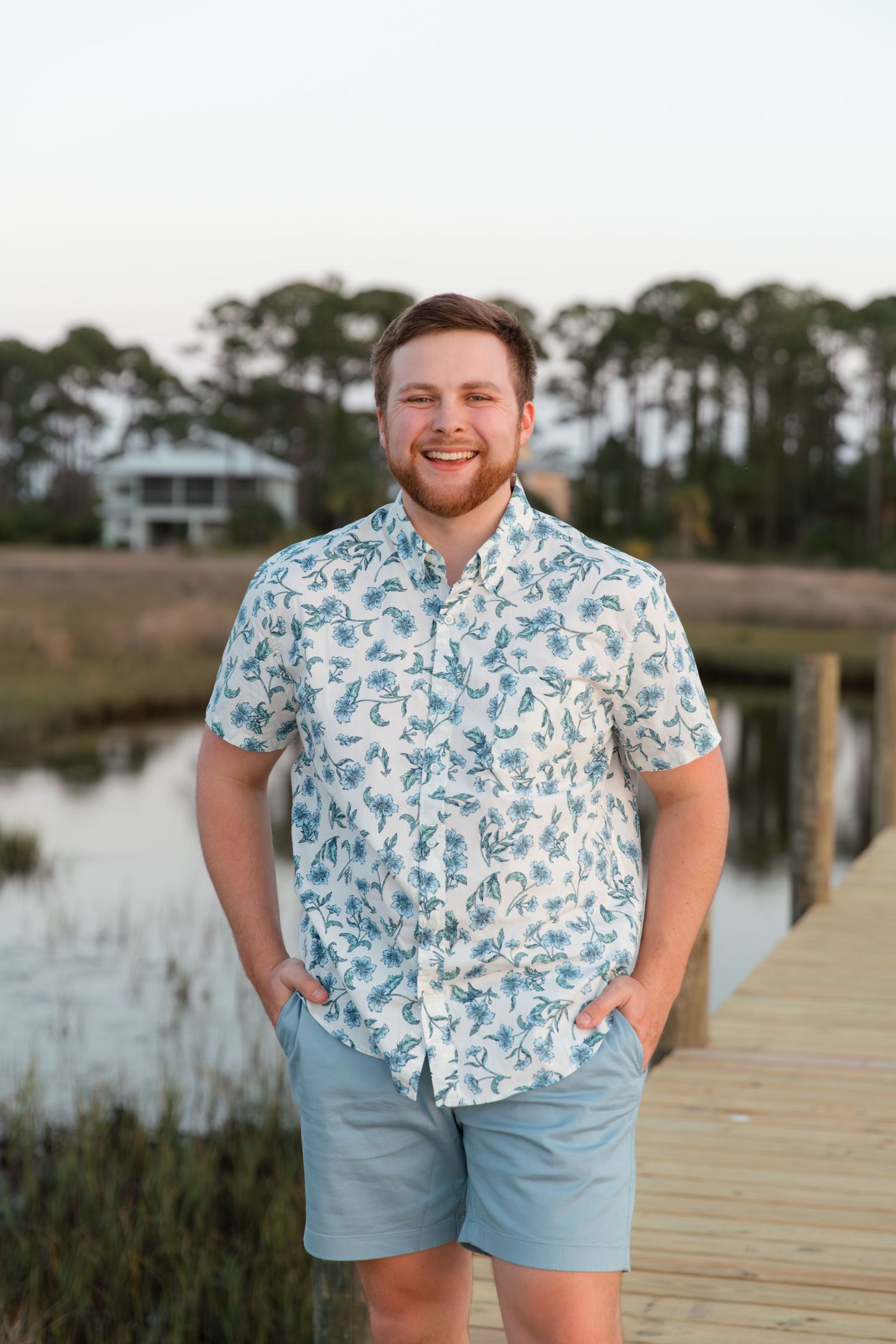 A person in a blue and white floral short-sleeve shirt and light blue shorts stands smiling on a wooden dock by a lake.