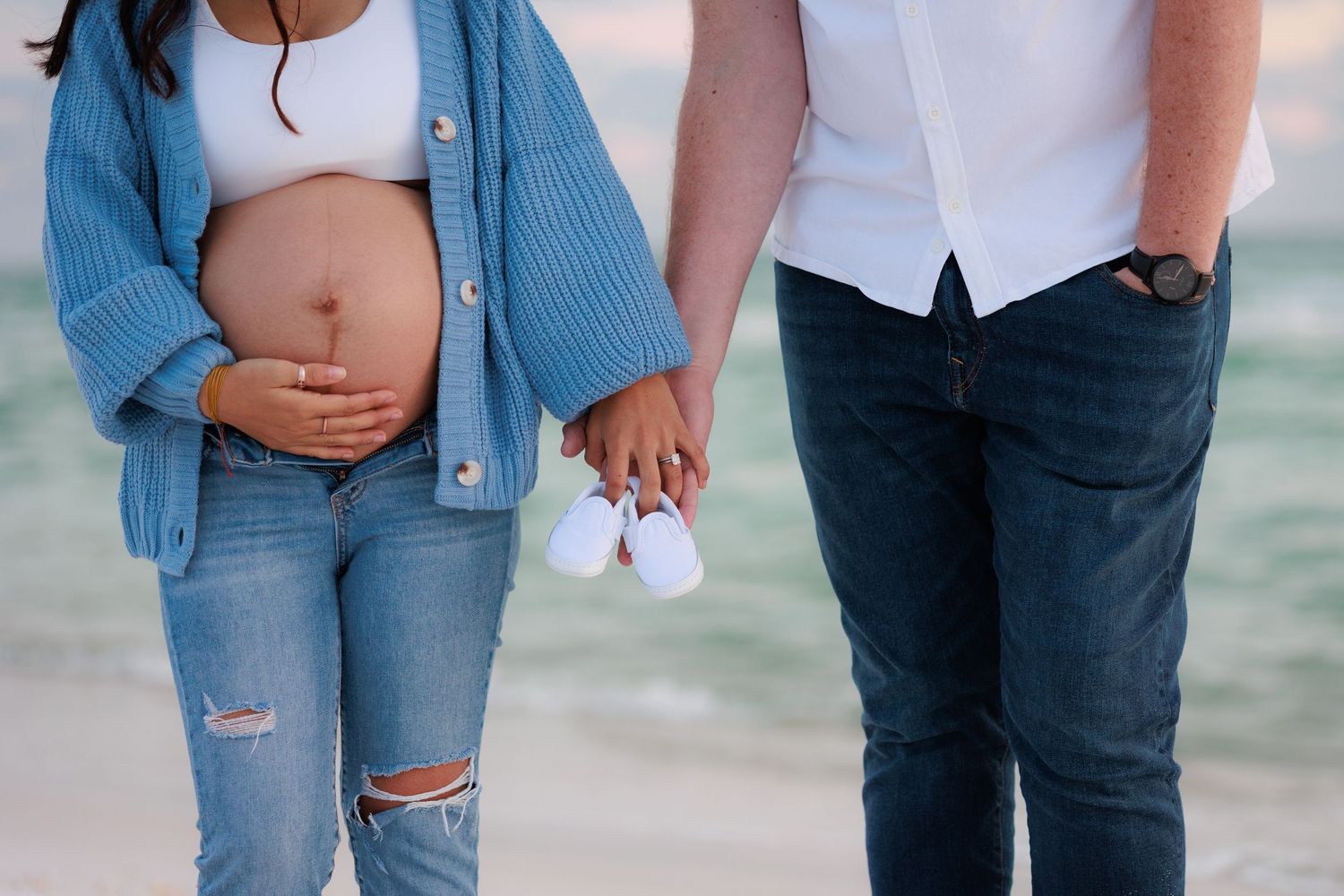 Close-up detail of expecting couple holding hands and baby shoes on the beach.
