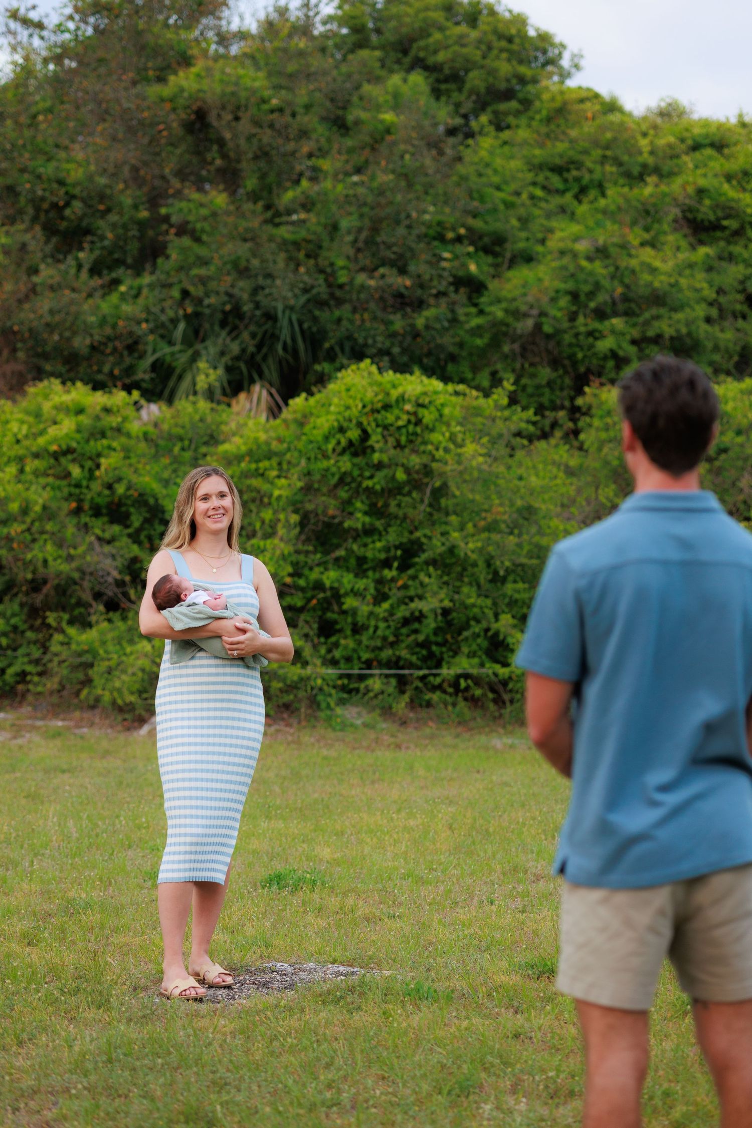 A couple poses together on a grassy hill surrounded by lush green trees during a sunny outdoor photoshoot.