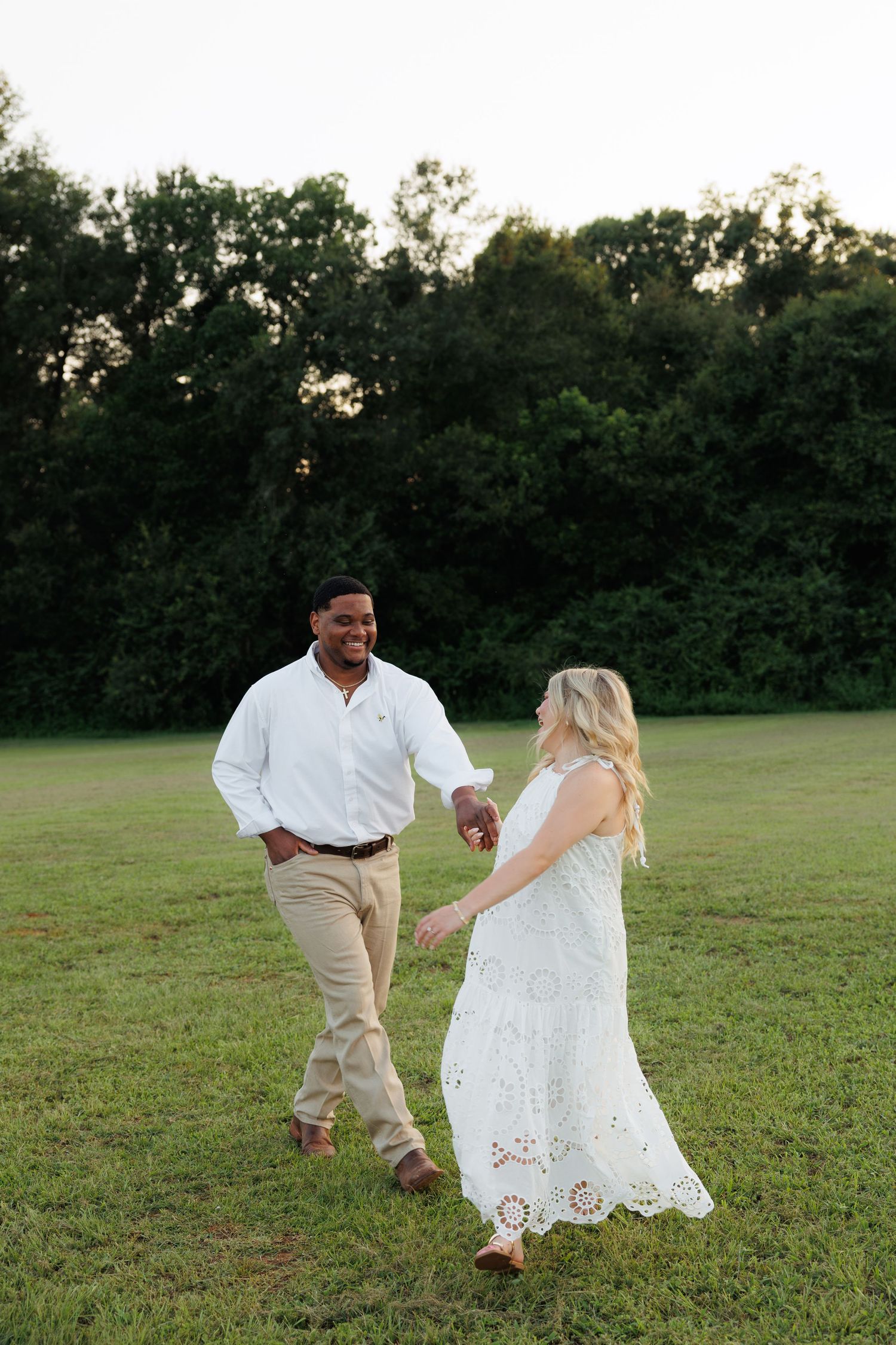 Smiling couple holding hands and walking together across a green field at golden hour.