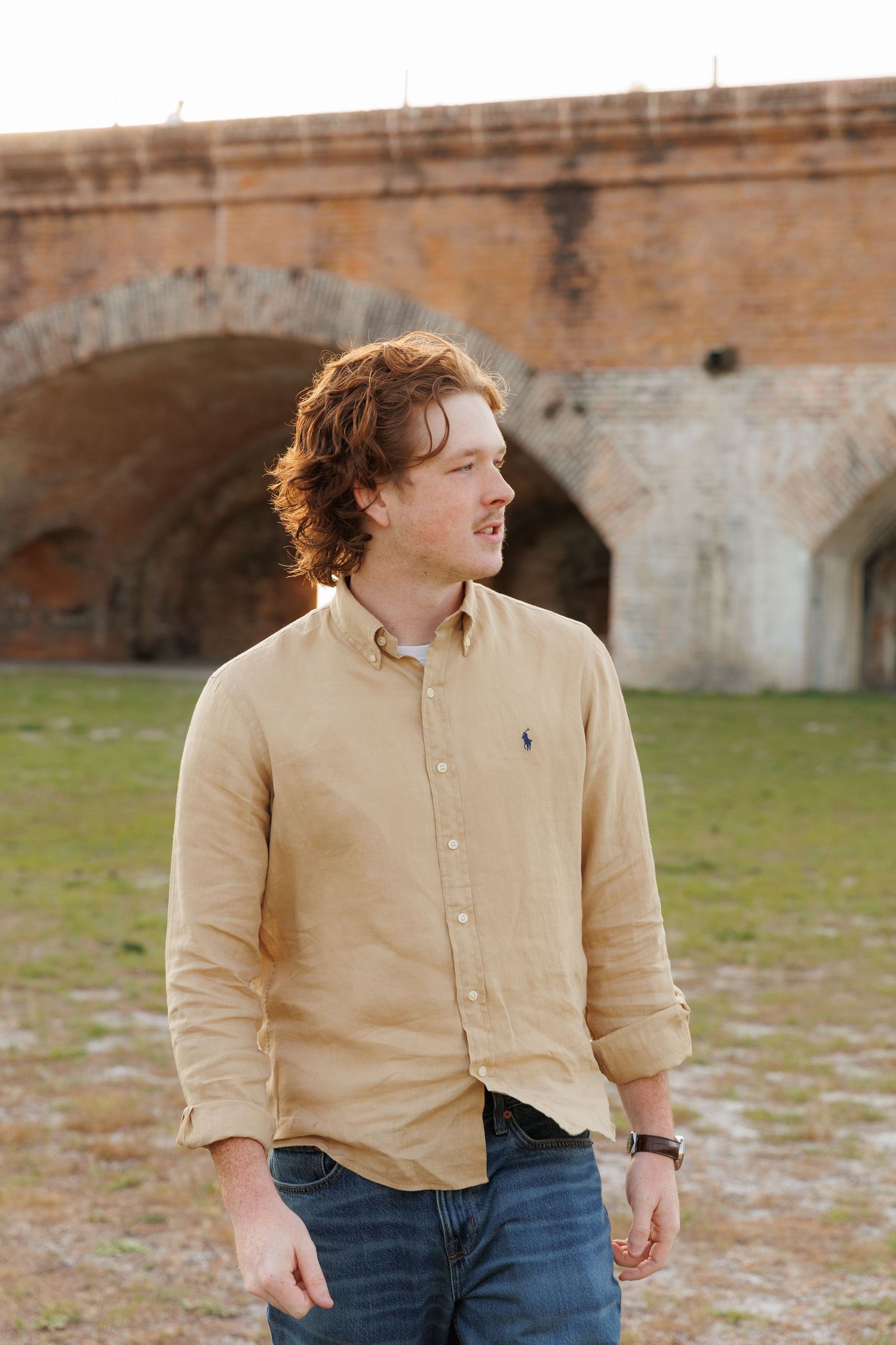 Young redhead man in tan Polo button-up shirt standing outdoors near rustic brick archway in natural light.
