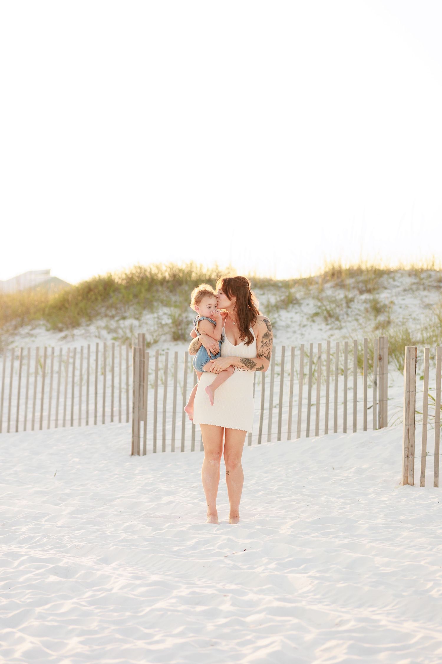 A mother holds and kisses her toddler on a white sand beach with dunes and a wooden fence backdrop.