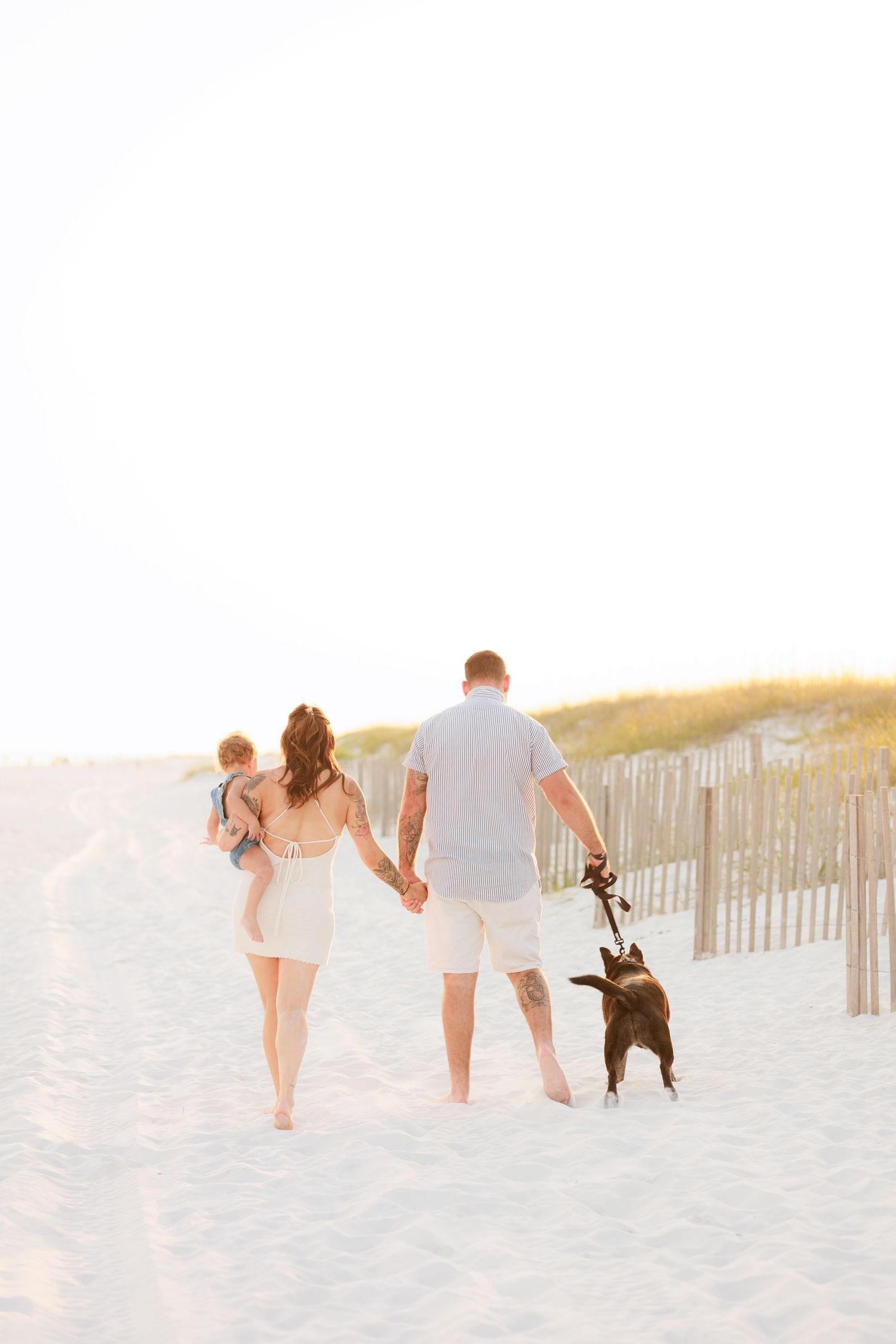 Family walking barefoot on white sand beach with dog and baby at sunset.