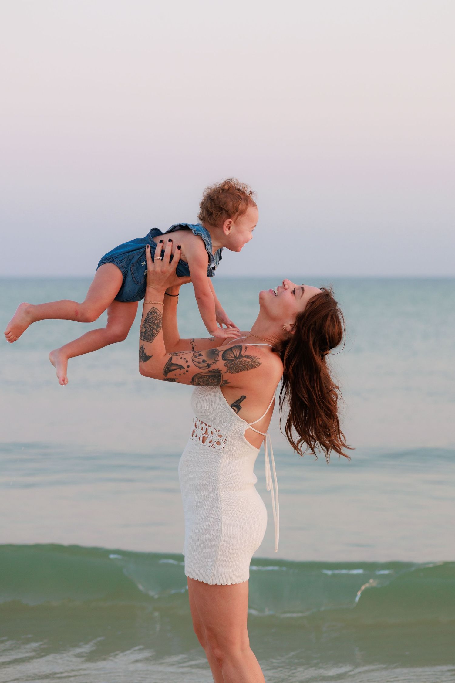 Mother lifting baby in air on beach at sunset, ocean behind them.