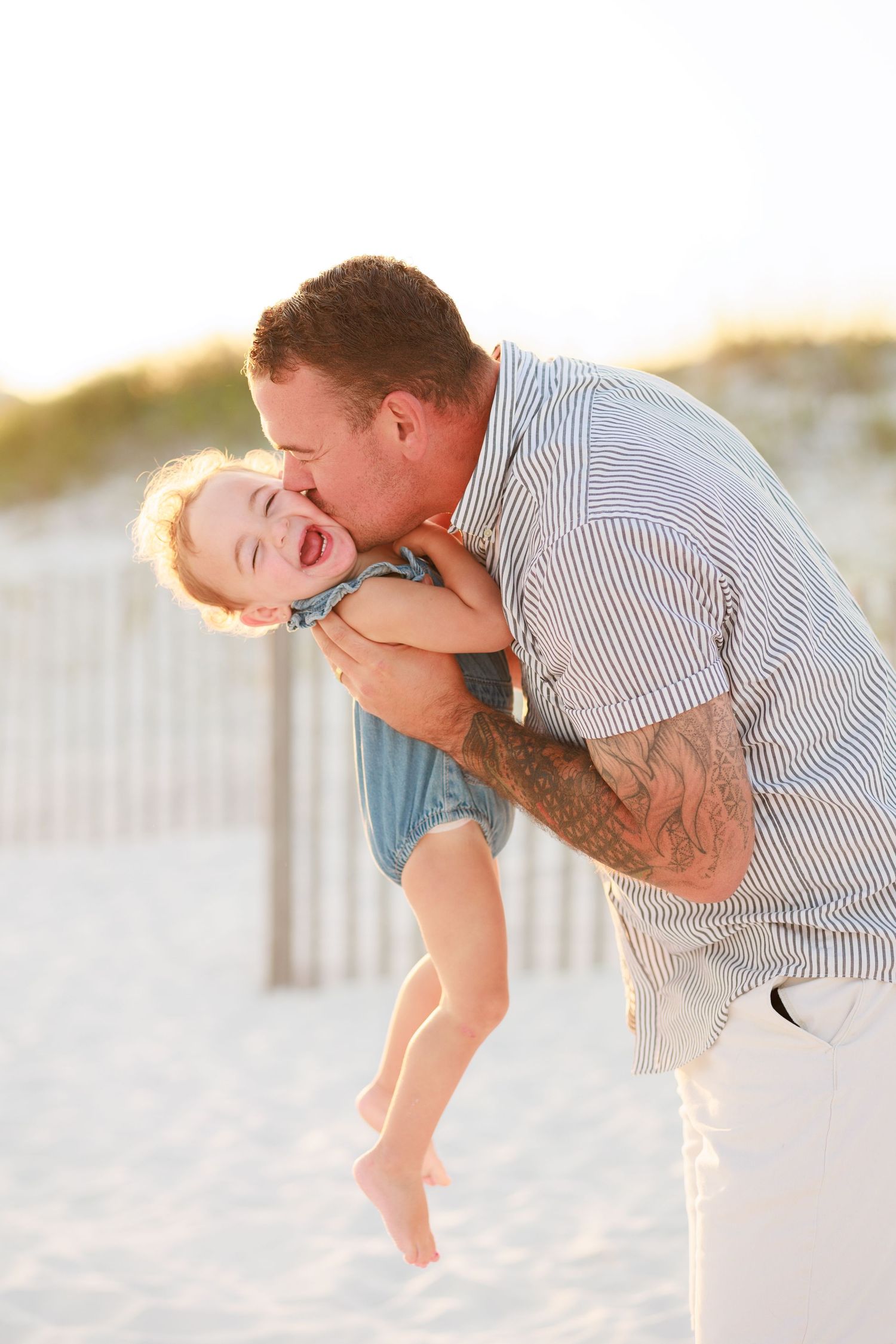 A father kisses his laughing toddler on a sunny beach, with sand dunes and a wooden fence in the background.