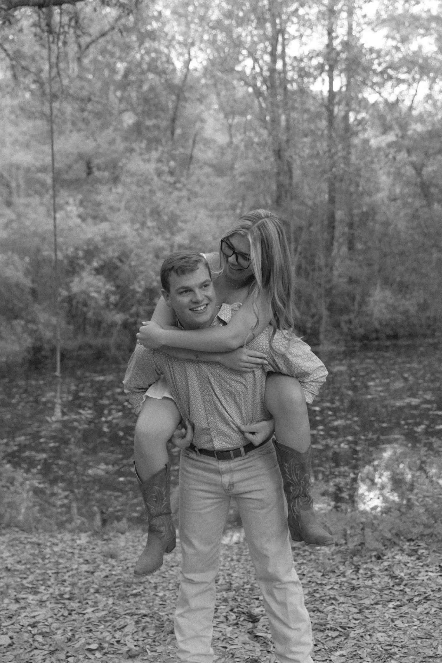 Black and white photo of a smiling man giving a woman a piggyback ride in a wooded outdoor setting.