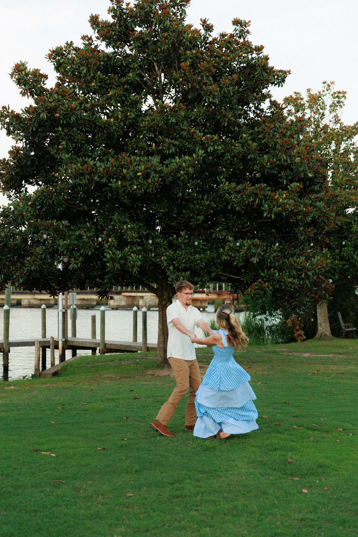 Couple twirling together on lush green grass near a scenic waterfront with a large magnolia tree backdrop.