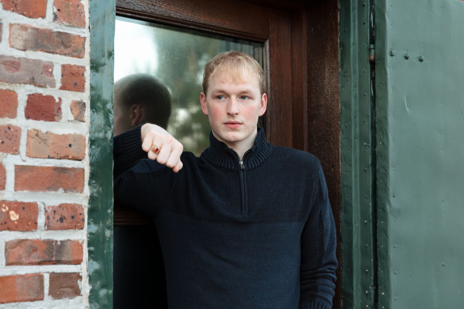 Young man in black zip-up sweater leaning against a green door frame beside a brick wall.
