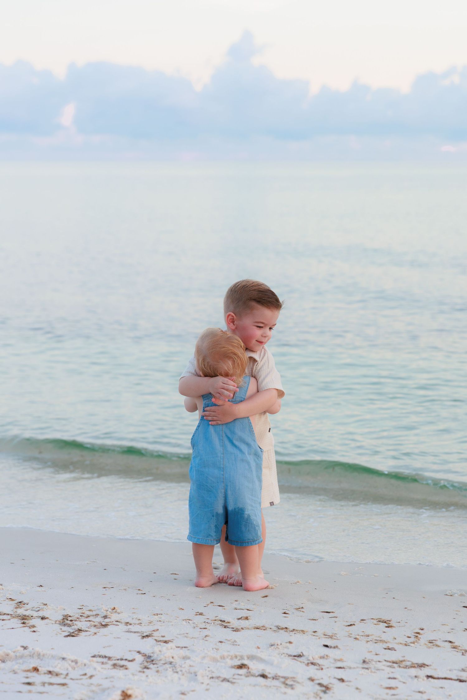 Two young siblings share a sweet hug on a white sandy beach with turquoise ocean waves in the background.