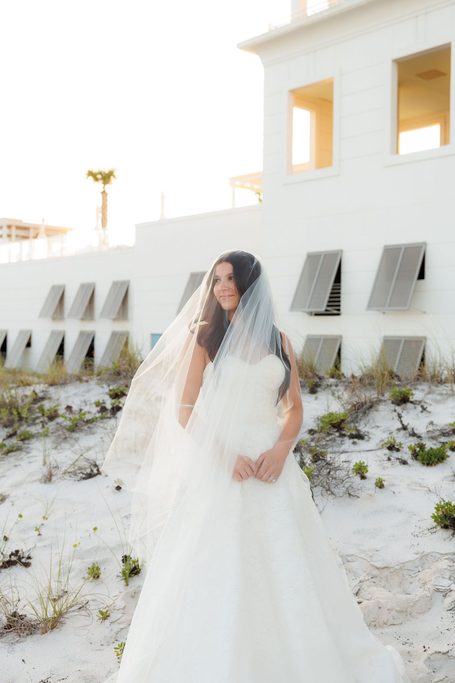 Bride in white strapless gown and veil poses on sandy beach dunes near white building at sunset.