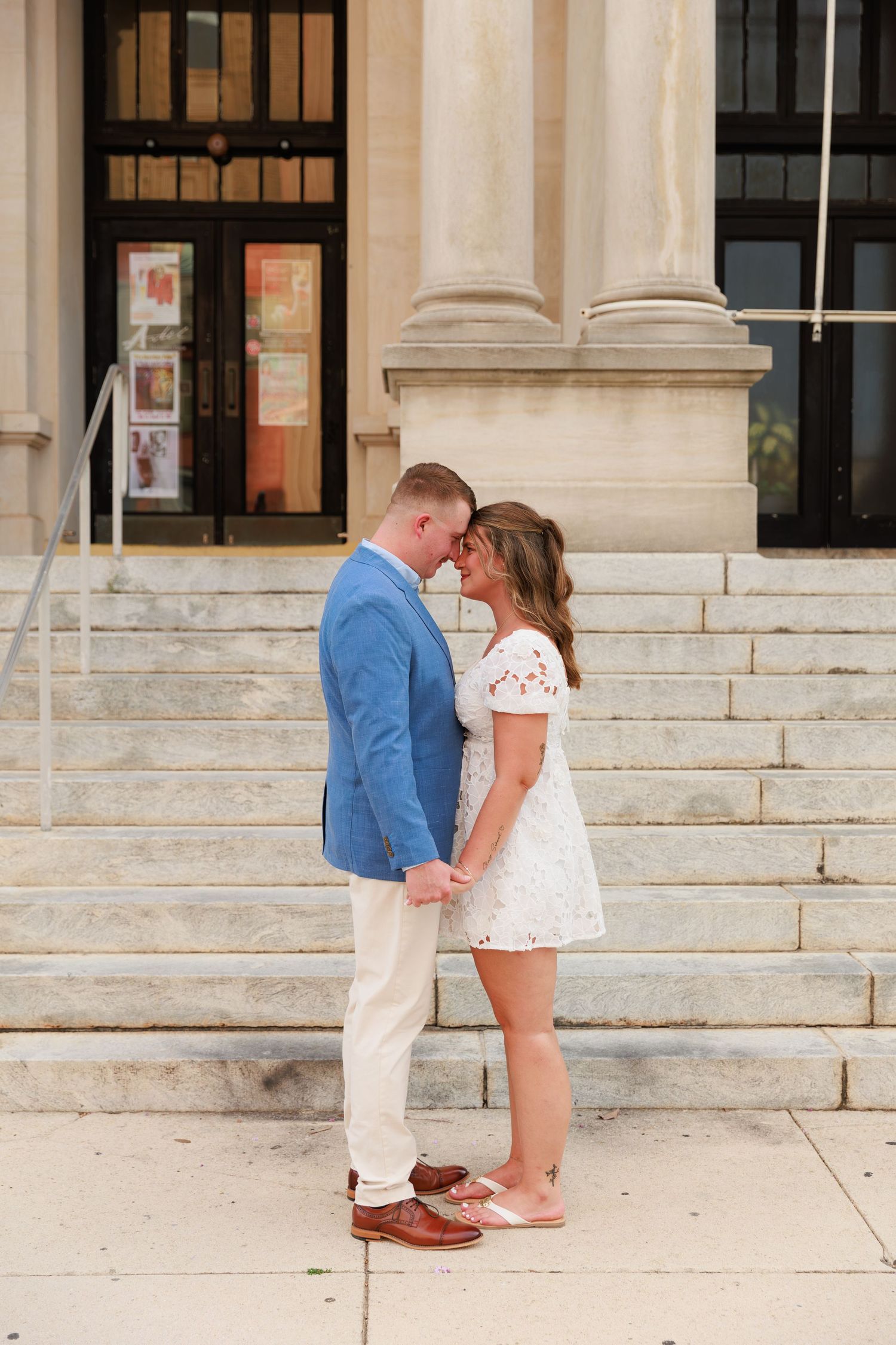 Engaged couple standing forehead to forehead holding hands in front of a grand stone building with columns.