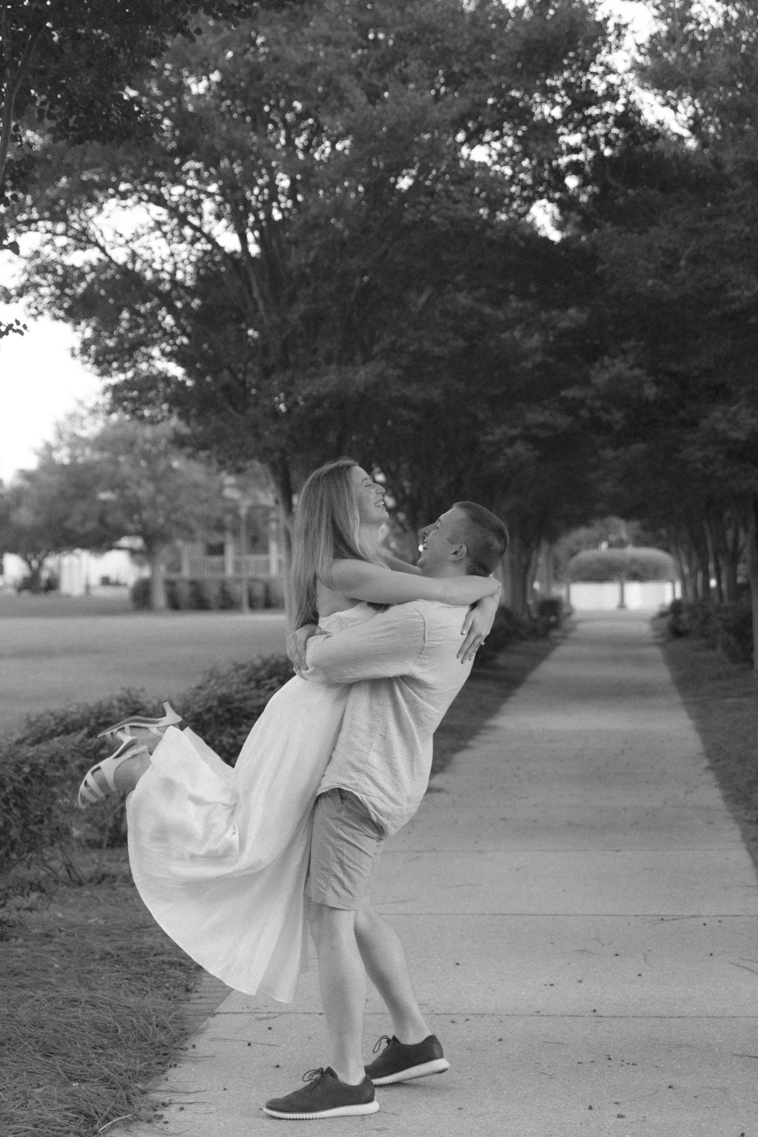 Black and white romantic photo of couple embracing on a tree-lined sidewalk, woman lifted off ground.