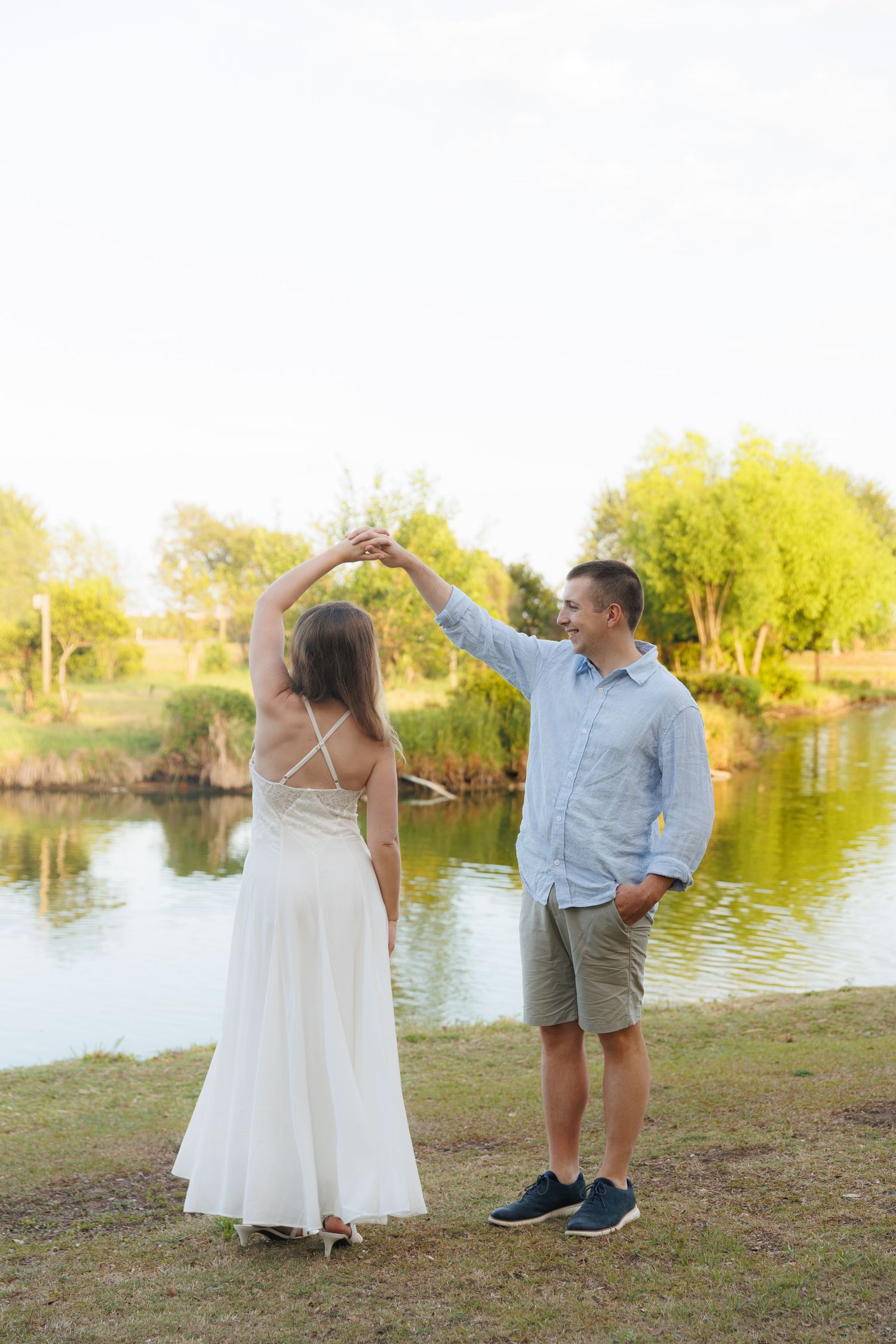 Couple dancing by a peaceful lake, man twirling woman in elegant white dress, surrounded by green trees at sunset.