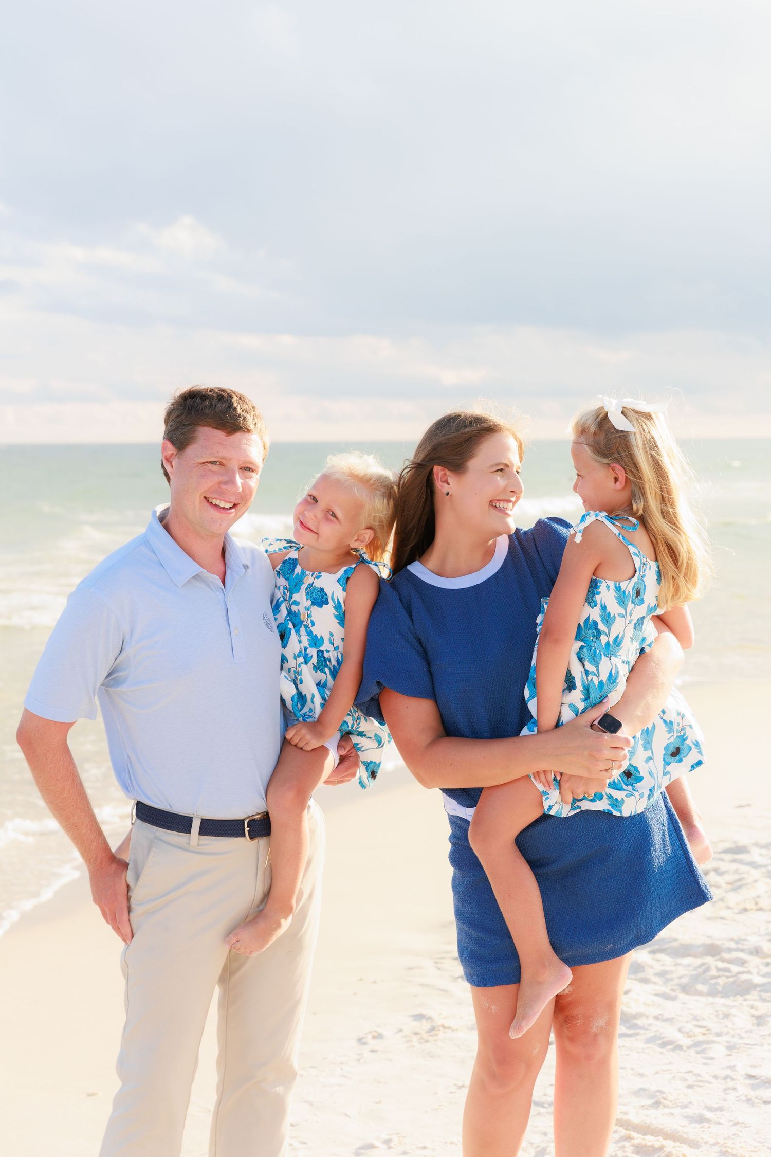 Family of four posing on a sunny beach, parents holding two young girls in matching blue floral dresses.