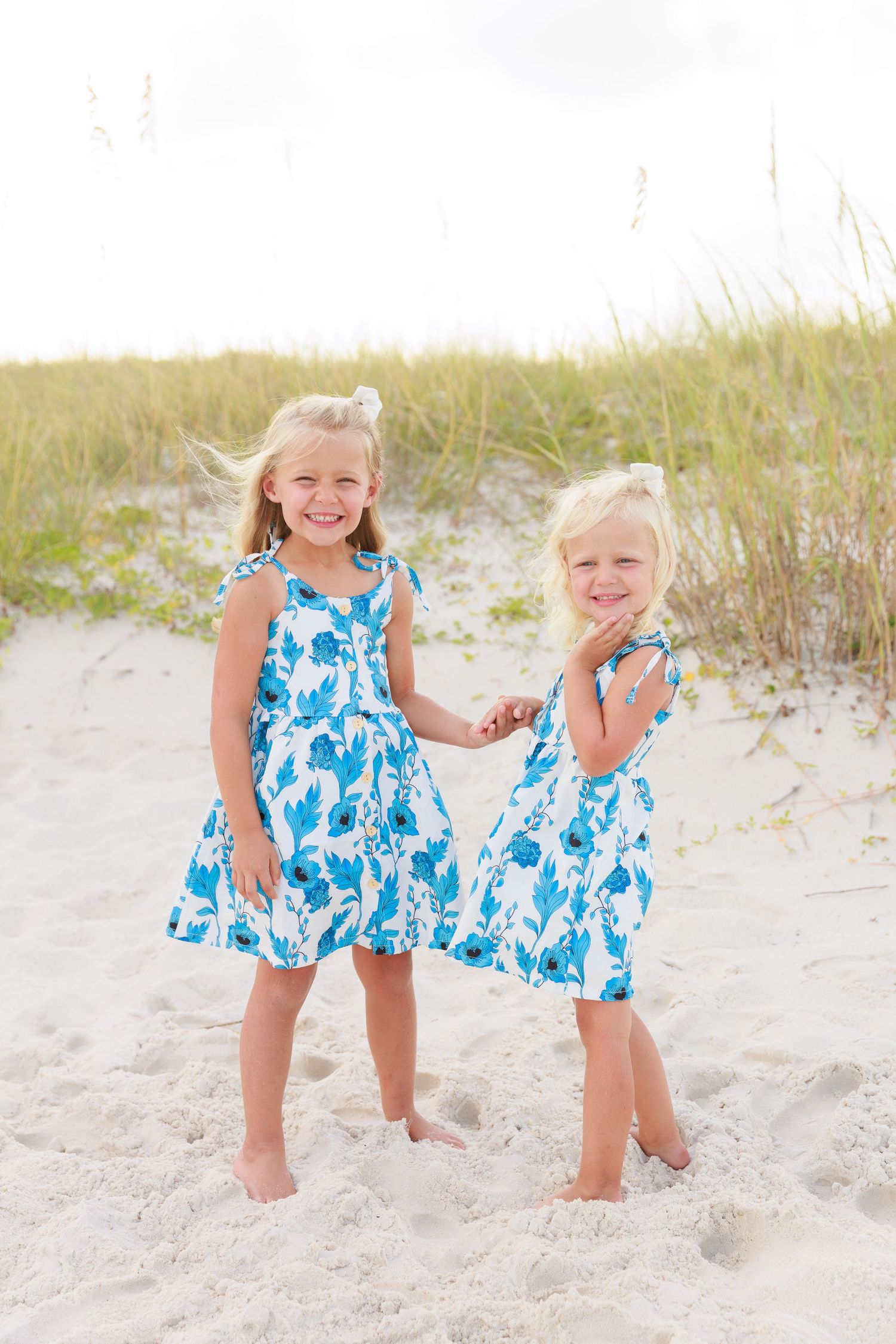 Two young girls in matching blue floral dresses smiling on a sandy beach with sea grass in the background.