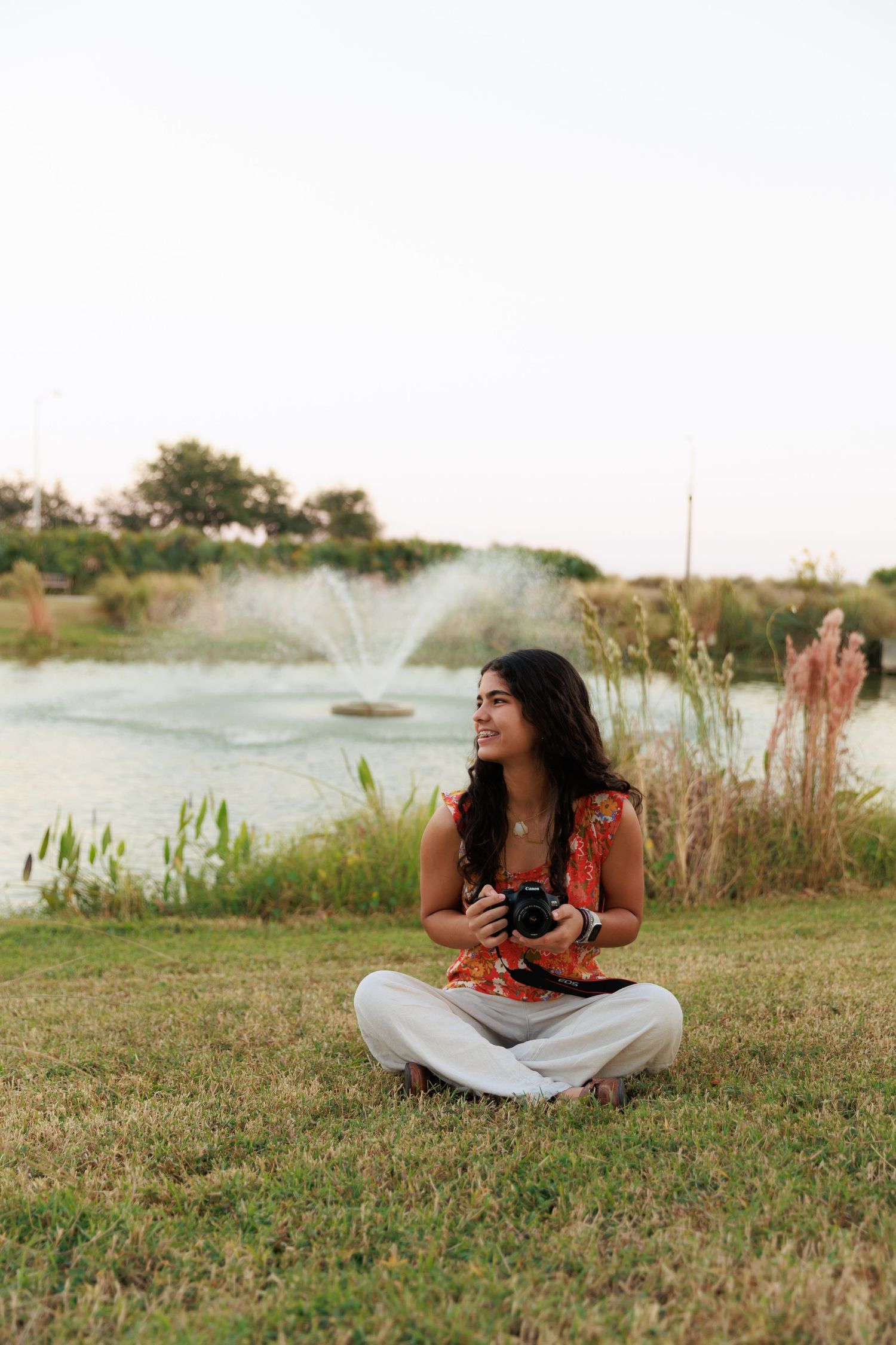 Nature photographer in casual attire sits by pond with fountain spray in background.